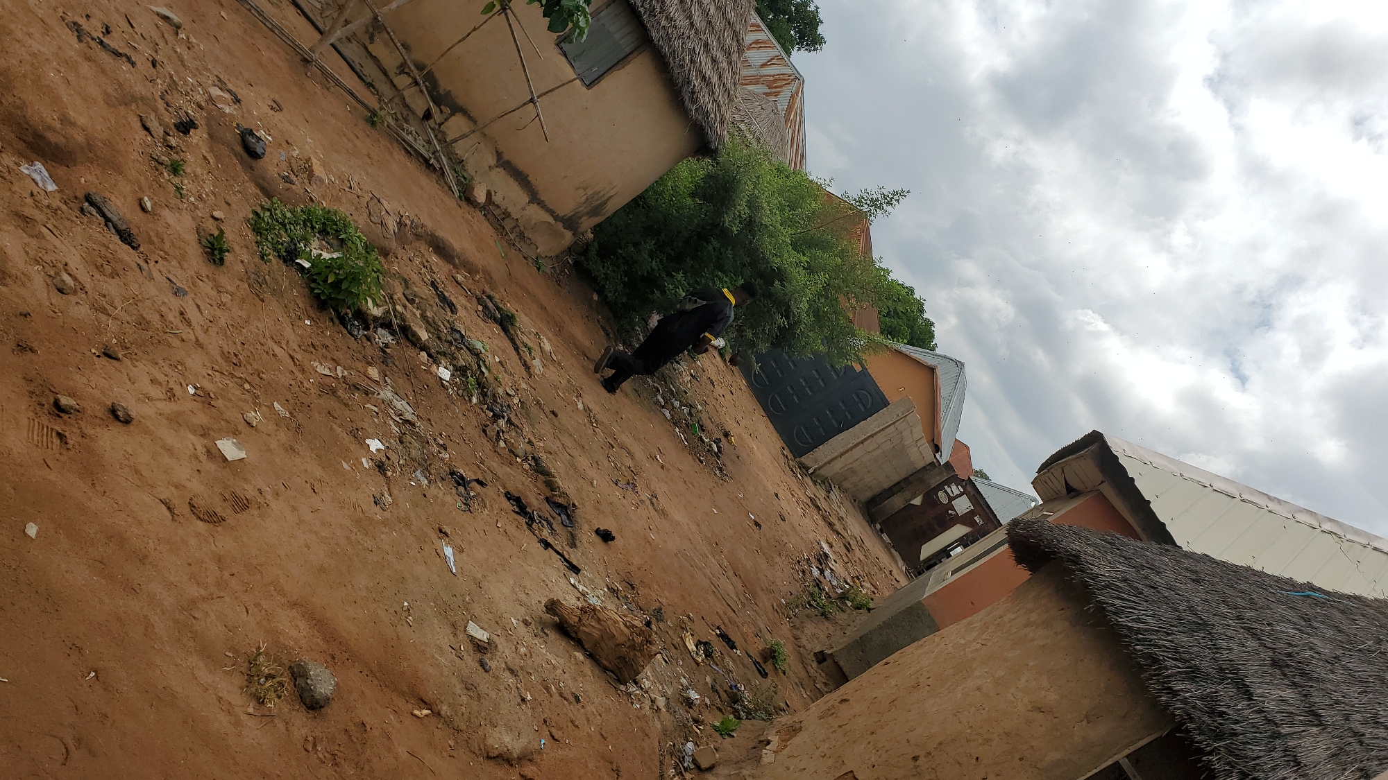 The image depicts a cluttered outdoor scene in the city of Jalingo, Nigeria. A person wearing a black garment with a yellow trim walks away from the camera in a dusty, unpaved area littered with debris. Buildings with thatched and corrugated metal roofs surround the area, some appearing to be made of mud. A large green bush is situated behind the person. The sky is overcast with scattered clouds.