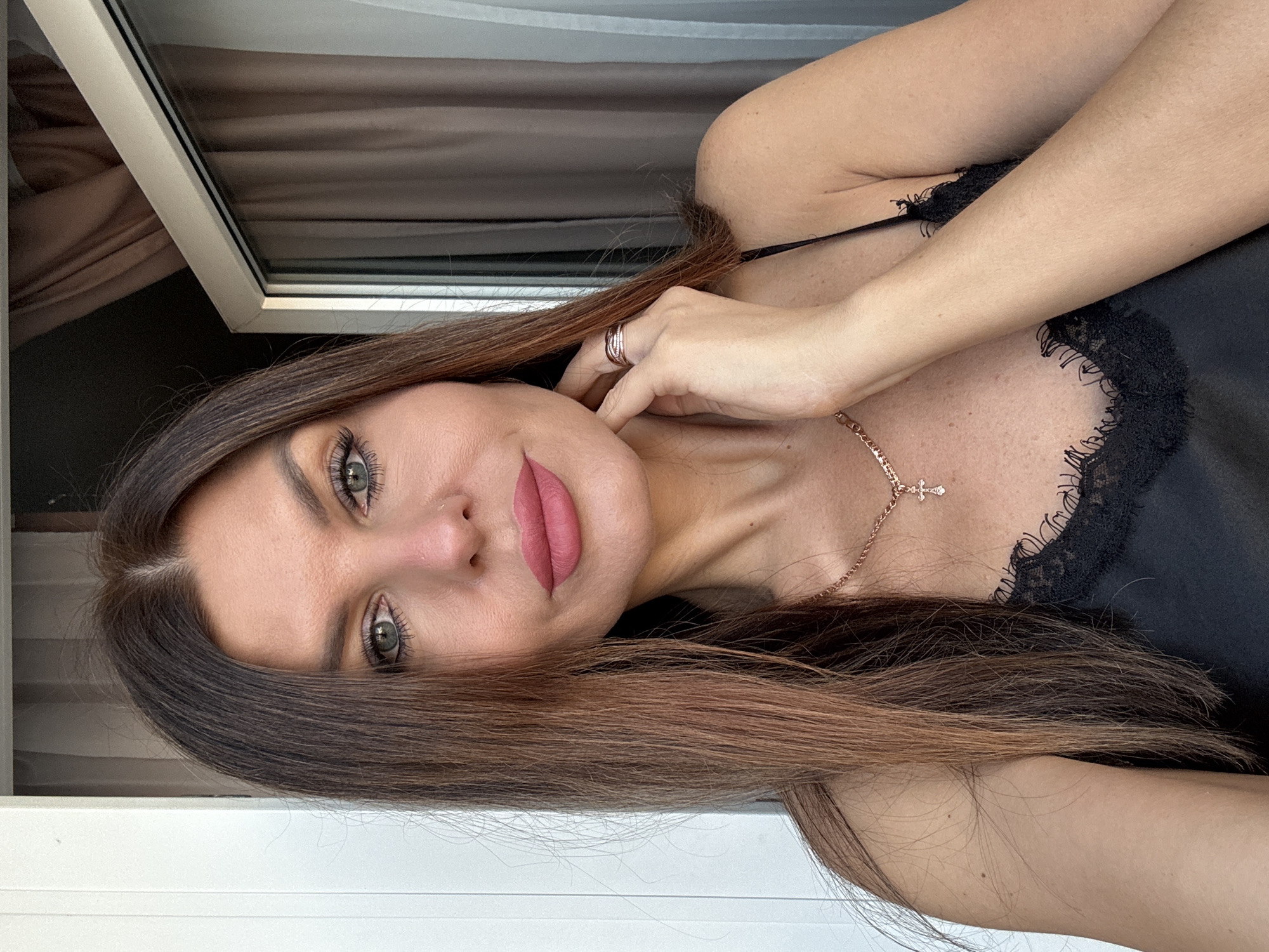 A fair-skinned woman with green eyes and dark brown hair poses indoors. She wears a black lace-trimmed top and a delicate gold necklace with a cross pendant. A ring is visible on her finger. A window frame and curtains are in the background. There is no verifiable information to confirm the location.