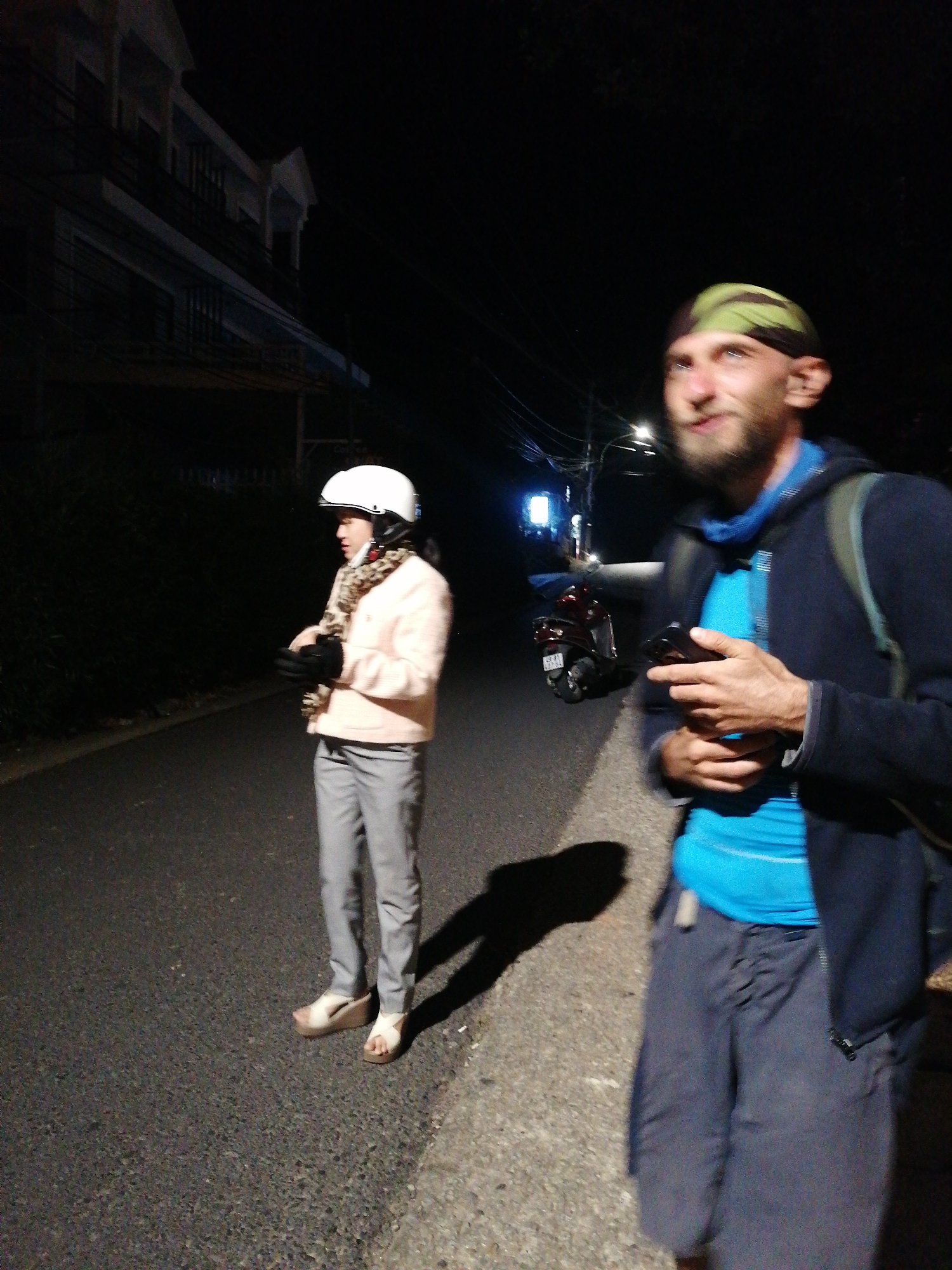 Two individuals are standing on an asphalt road at night. On the left, a person wearing a white helmet, a light pink jacket, a patterned scarf, and gray pants stands facing to the right. They are wearing white wedge sandals and black gloves. On the right, a person with a beard and a green and black bandana is looking up and to the left, holding a dark-colored mobile phone in their hands. They are wearing a blue shirt, a dark blue jacket, and dark pants, with a backpack slung over their left shoulder. A red scooter is parked on the left side of the road behind the first individual. A building with multiple balconies is visible in the background on the left. Streetlights and power lines are present in the background. The scene appears to be a street in Da Lat, Vietnam, indicated by the presence of a scooter commonly used in the region and the architectural style of the building.