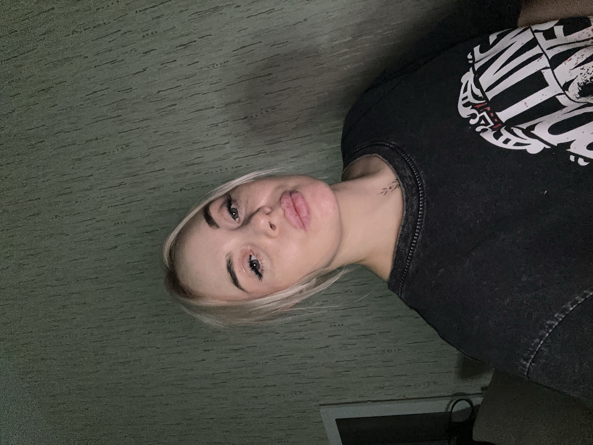 A young woman with blonde hair is visible. She has light skin and is wearing a dark shirt with white graphics. A small tattoo is visible on her neck. She is indoors, against a textured wall with a green hue. The location is Vysokiy, Russia.