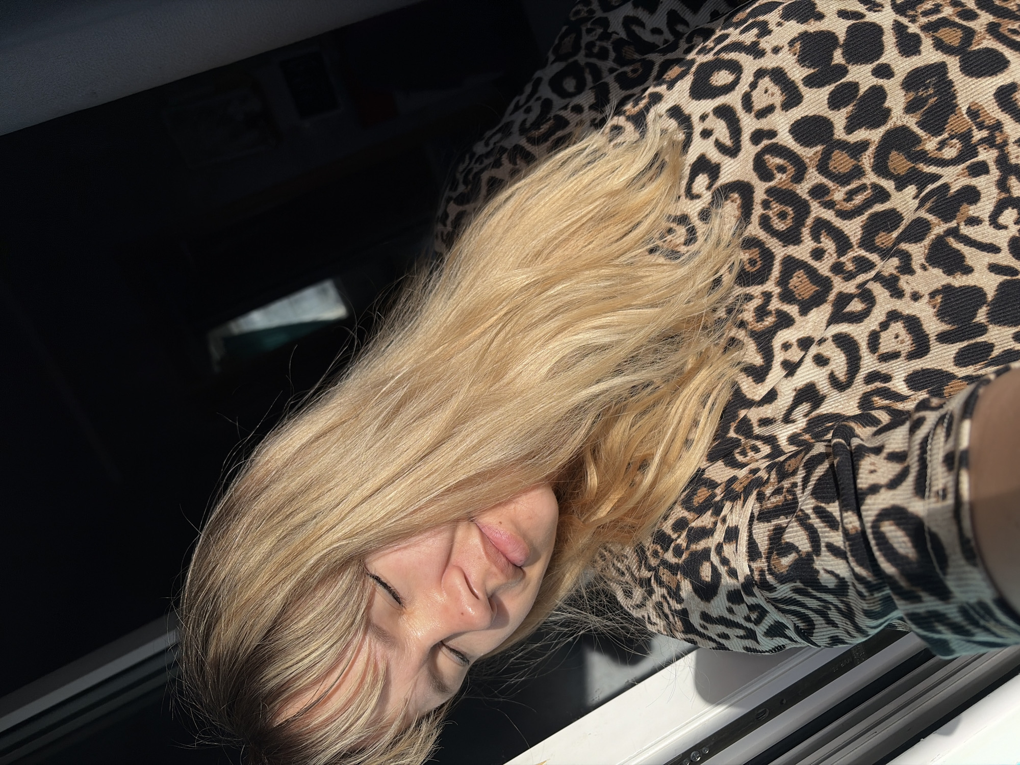 A blonde woman wearing a leopard print shirt is sitting by a window with black frame parts. The location is Chaikovsky, Russia.