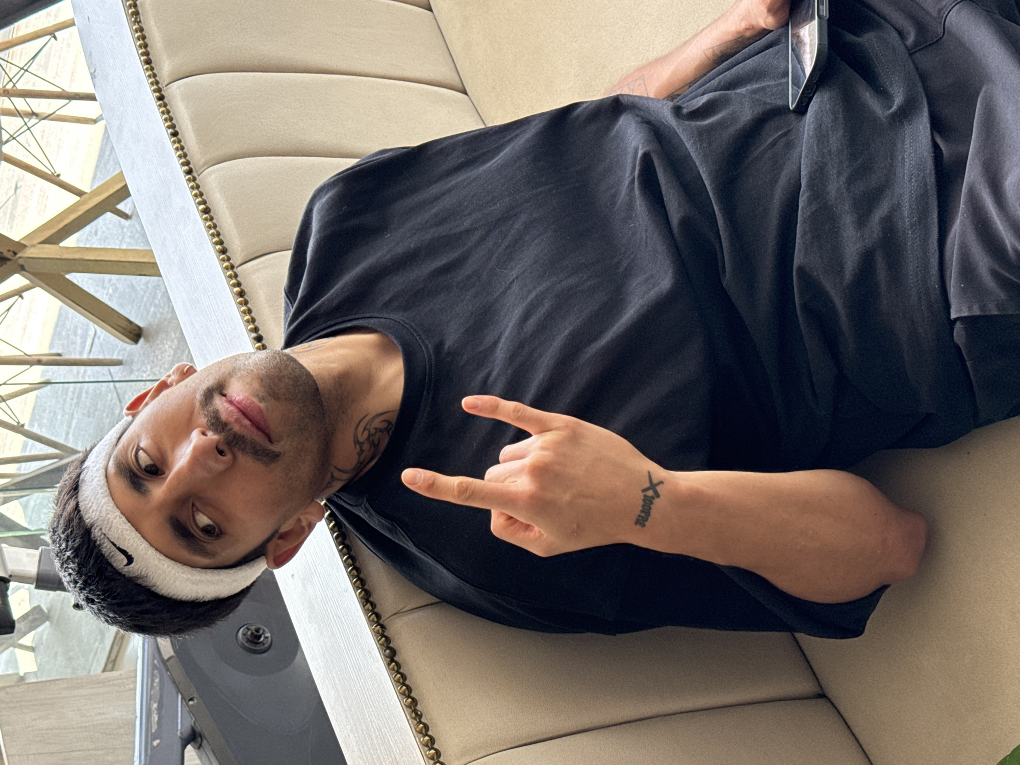 A man is captured lying down on a light-colored, upholstered sofa in an indoor setting, likely a contemporary living room or lounge area in Puebla, Mexico. He is wearing a black short-sleeved t-shirt and a white Nike headband. His head is slightly tilted, and he looks directly at the camera with a neutral to slightly serious expression. He has a mustache and a short beard, along with a tattoo visible on his neck/shoulder area.
His right hand is raised, forming a "rock on" or "devil horns" gesture, with a tattoo reading "Xaverne" clearly visible on his right wrist. Another tattoo is visible on his left arm, which is resting along his side. A smartphone, with a transparent case, is tucked into the left side of his black t-shirt. The sofa features a decorative gold stud trim along its edge. In the background, bright lighting suggests it is daytime, illuminating modern furniture elements, including what appears to be a table with a geometric, possibly metallic, base.