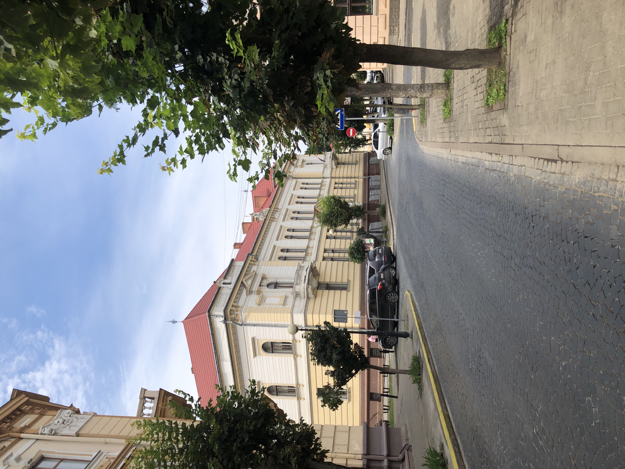 The image depicts a street scene in Kolomyia, Ukraine. A two-story building with a red tiled roof and cream-colored facade is visible on the left. The building has arched windows and decorative architectural elements. In front of the building, there are trees and a cobblestone street. Several cars are parked along the curb. The sidewalk is made of paving stones, and more trees are planted along it. The sky is partly cloudy. The scene suggests a quiet urban environment, possibly a historical district, given the architecture and cobblestone road.