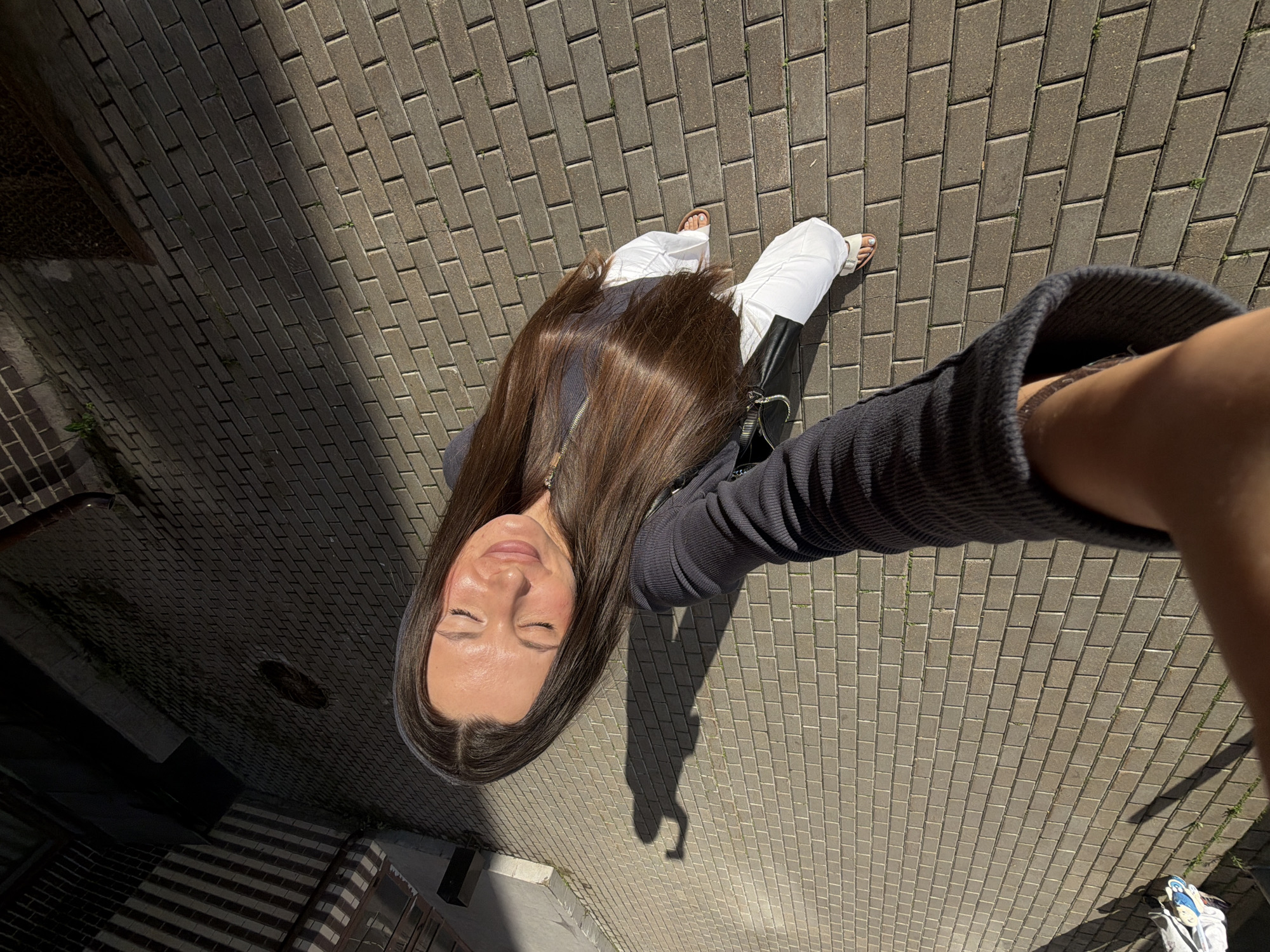 A woman with long brown hair is taking a selfie outdoors on a paved area in Odintsovo, Russia. She is wearing a dark long-sleeved top and white pants. Her eyes are closed and her face is tilted up towards the sun, suggesting she is enjoying the warmth. The ground is made of rectangular bricks, and there is a shadow of the woman cast on the bricks. In the background, there are some structures and a fence. The overall scene is casual and relaxed.