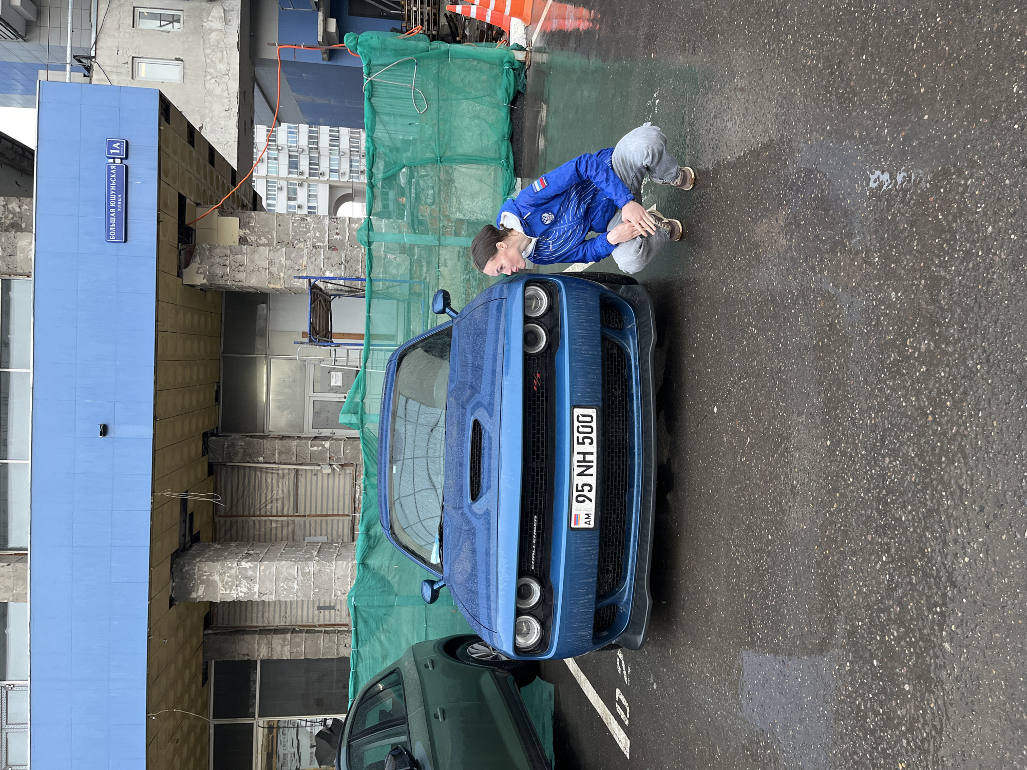 A woman squats in front of a blue Dodge Challenger in Moscow, Russia on a wet day. The woman has dark hair and is wearing a blue jacket with a Russian flag patch on the sleeve, gray pants, and brown shoes. The car has a license plate reading "95 NH 500" with the Armenian flag displayed to the left of the license plate number. The location is identifiable by a street sign written in Cyrillic, showing "Большая Юшуньская улица" and "1А" (Bolshaya Yushunskaya Street, 1A). The scene includes construction material such as green netting and scaffolding around a building in the background. Another, dark-colored vehicle is partially visible in the lower left corner of the frame.