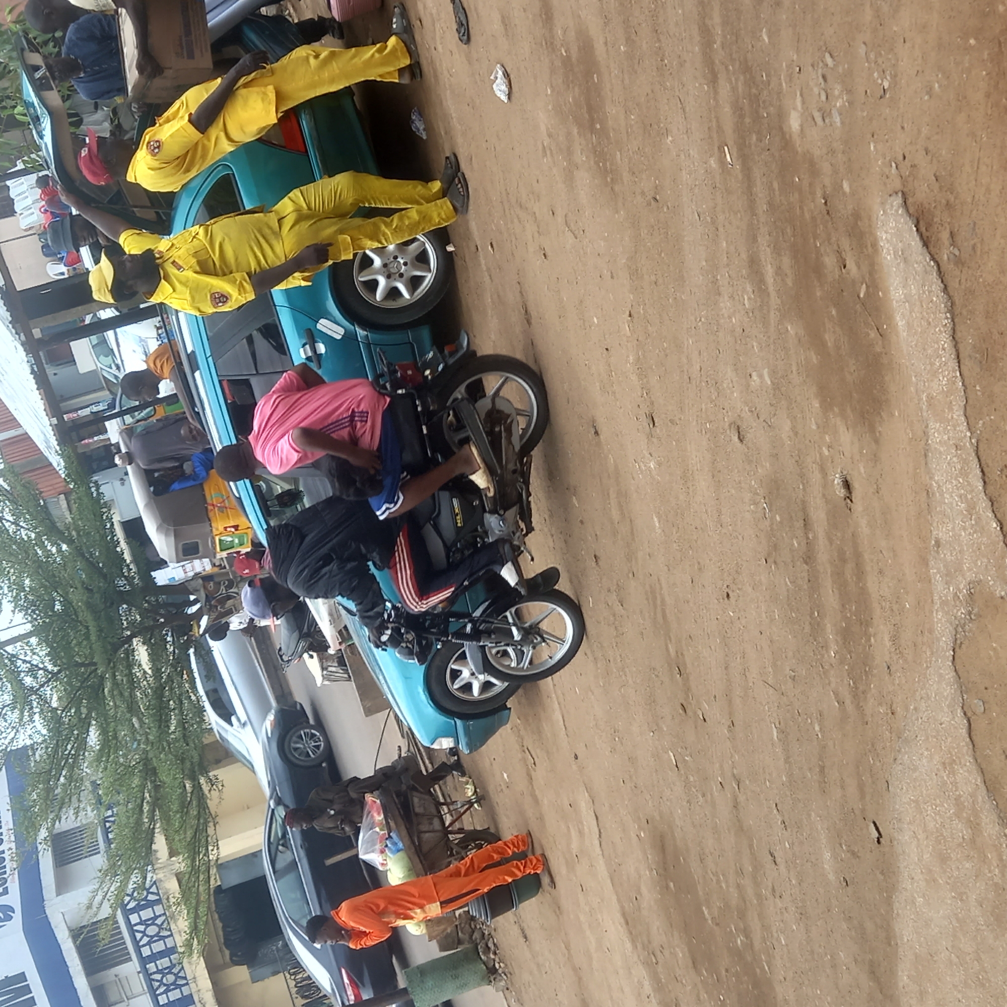 In Dadi Riba, Nigeria, a teal-colored car is parked on a dirt street. Several individuals are interacting around the car. Two men wearing yellow uniforms are positioned on top of the car's roof. A man in a pink shirt is near a motorcycle that is partially supported by the car. A man wearing an orange jumpsuit is standing near the rear of a black car, and there are other individuals present. A multi-level building is visible in the background. The scene appears to be taken outdoors.