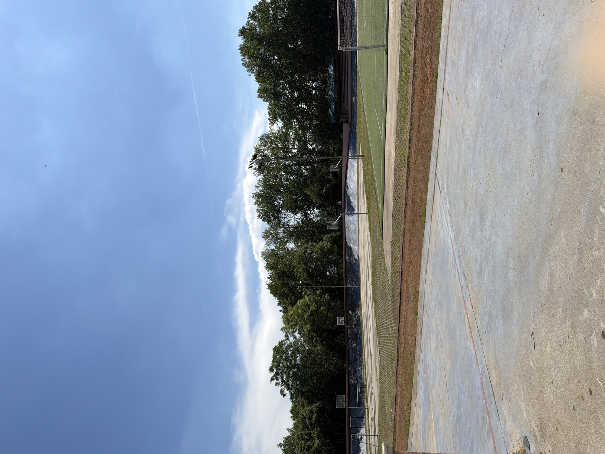 The image shows an outdoor sports complex under a bright blue sky with scattered white clouds. In the foreground, a concrete court with red markings is visible. Beyond this, a paved area with a grid pattern separates it from a green field, likely a soccer pitch, which features a white goal net on the right side. A line of trees forms the backdrop, with some smaller structures and basketball hoops interspersed among them. The overall impression is of a public recreational facility.