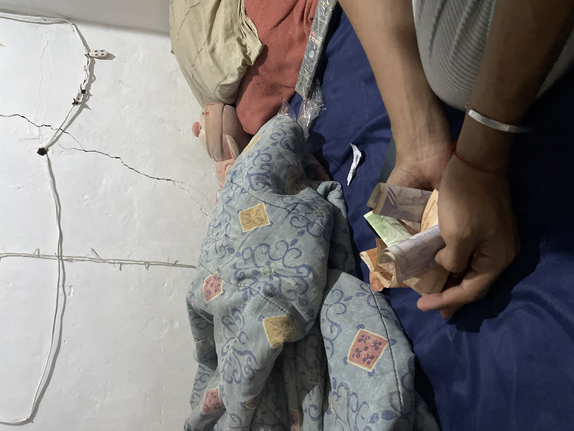 The image depicts a close-up view of a person's hands holding a bundle of folded banknotes. The hands are positioned over a dark blue surface, likely a bed or couch, which is partially covered by a patterned blue and grey blanket. In the background, a white wall is visible, marked with a prominent crack and a series of electrical wires and a power outlet. A remote control is placed on a red blanket to the right of the folded money. The lighting suggests an indoor environment. The specific location within the city of Unknown, Unknown, cannot be determined from the visual information provided.