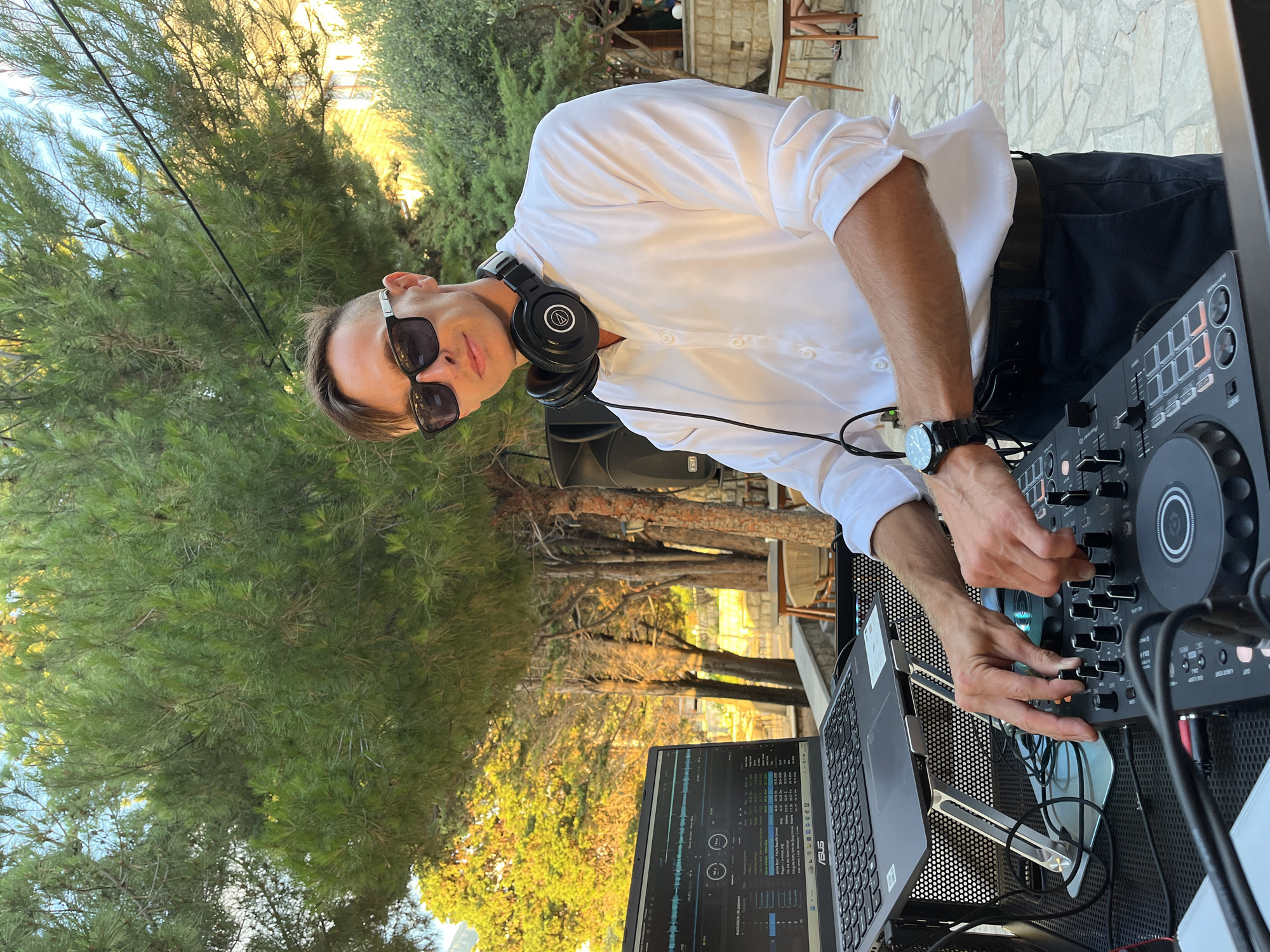 A male DJ, wearing sunglasses and a white collared shirt, stands behind a DJ setup outdoors. He has headphones around his neck and is operating a DJ controller. In front of him is a laptop displaying music software, positioned on a stand. The DJ setup includes speakers. The background shows trees, with yellow-leafed trees on the left, suggesting autumn. In the far background, tables and chairs are visible, and some structures. The ground is paved with stone. The location is identified as Pečurice, Montenegro.