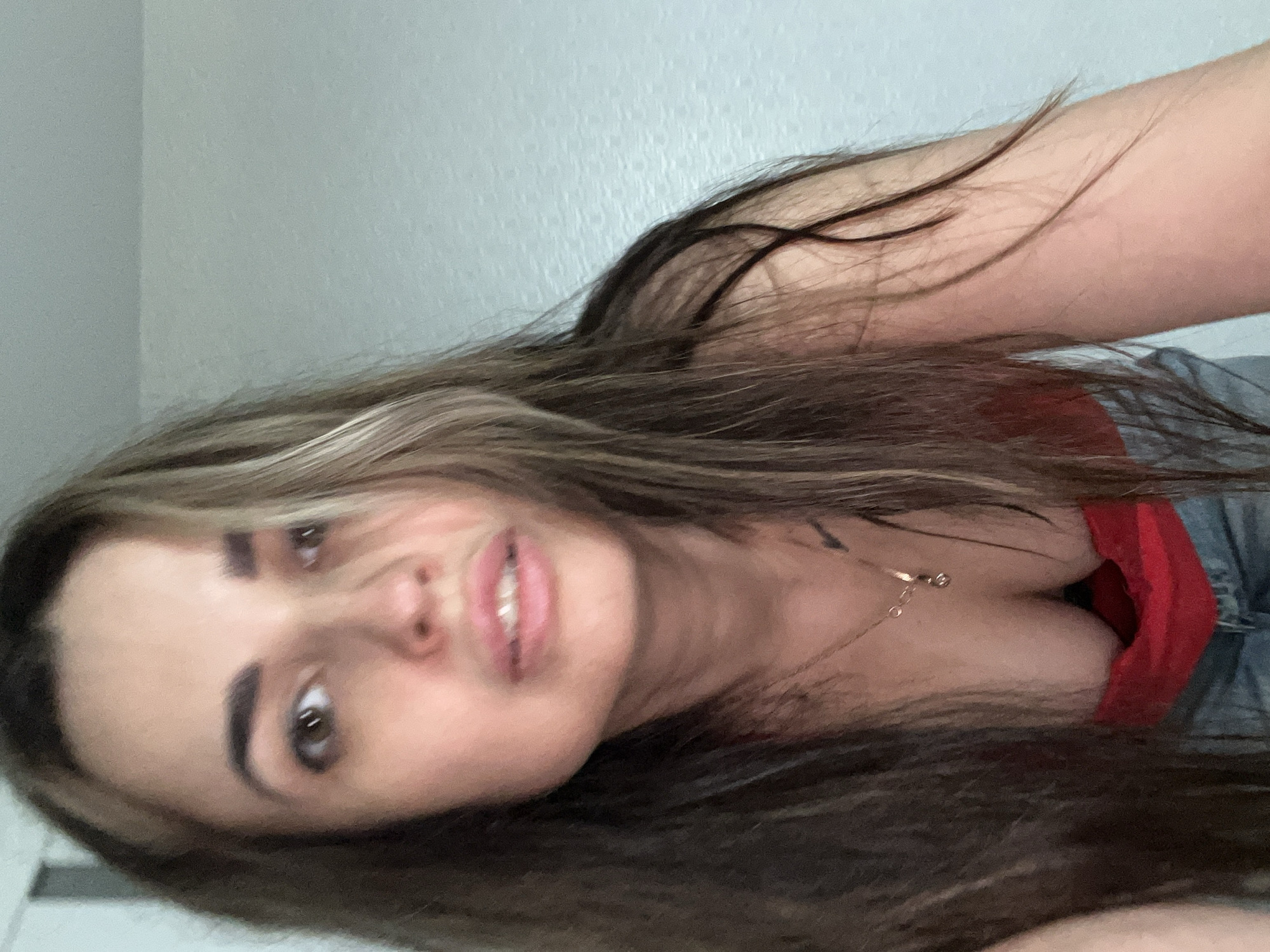 A close-up, rotated portrait shows a young woman looking directly at the camera. Her head is tilted, and the image itself is oriented horizontally, with her head on the left and her body extending to the right. She has long, dark brown hair with lighter highlights, which cascades around her face and extends across the bottom of the frame. Her right eye is clearly visible, dark brown, and looking upwards (towards the top of the frame in the current orientation). Her lips are slightly parted in a soft expression, revealing a hint of teeth. She has well-defined eyebrows.
She is wearing a red top with a low neckline, partially visible on the right side of the frame, and a light blue denim garment (possibly jeans or shorts) is also visible. A delicate gold-colored necklace with a small pendant is around her neck, resting on her chest. The background appears to be a plain, light-colored wall or surface. The lighting is soft and even, suggesting an indoor setting. There is no visible text. The location is unknown.