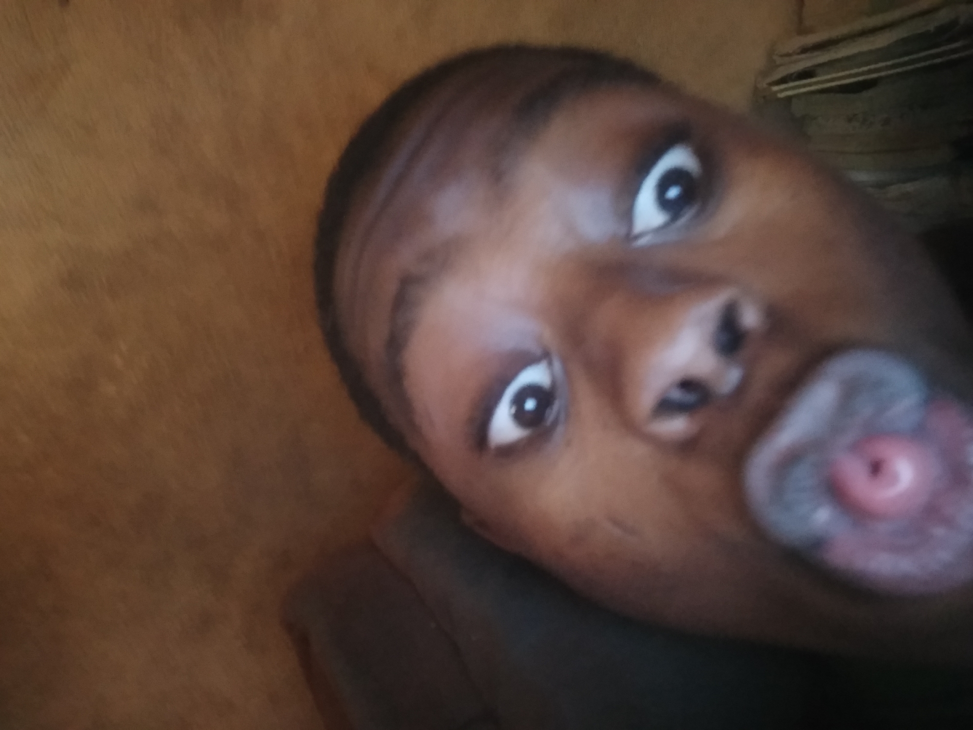 A close-up photograph depicts a person with dark skin, wide eyes, and a partially open mouth with their tongue extended and rolled. The background appears to be an indoor setting with a textured wall partially visible. The location is noted as Shangisha, Nigeria, but there is no information in the picture to verify the exact surroundings.