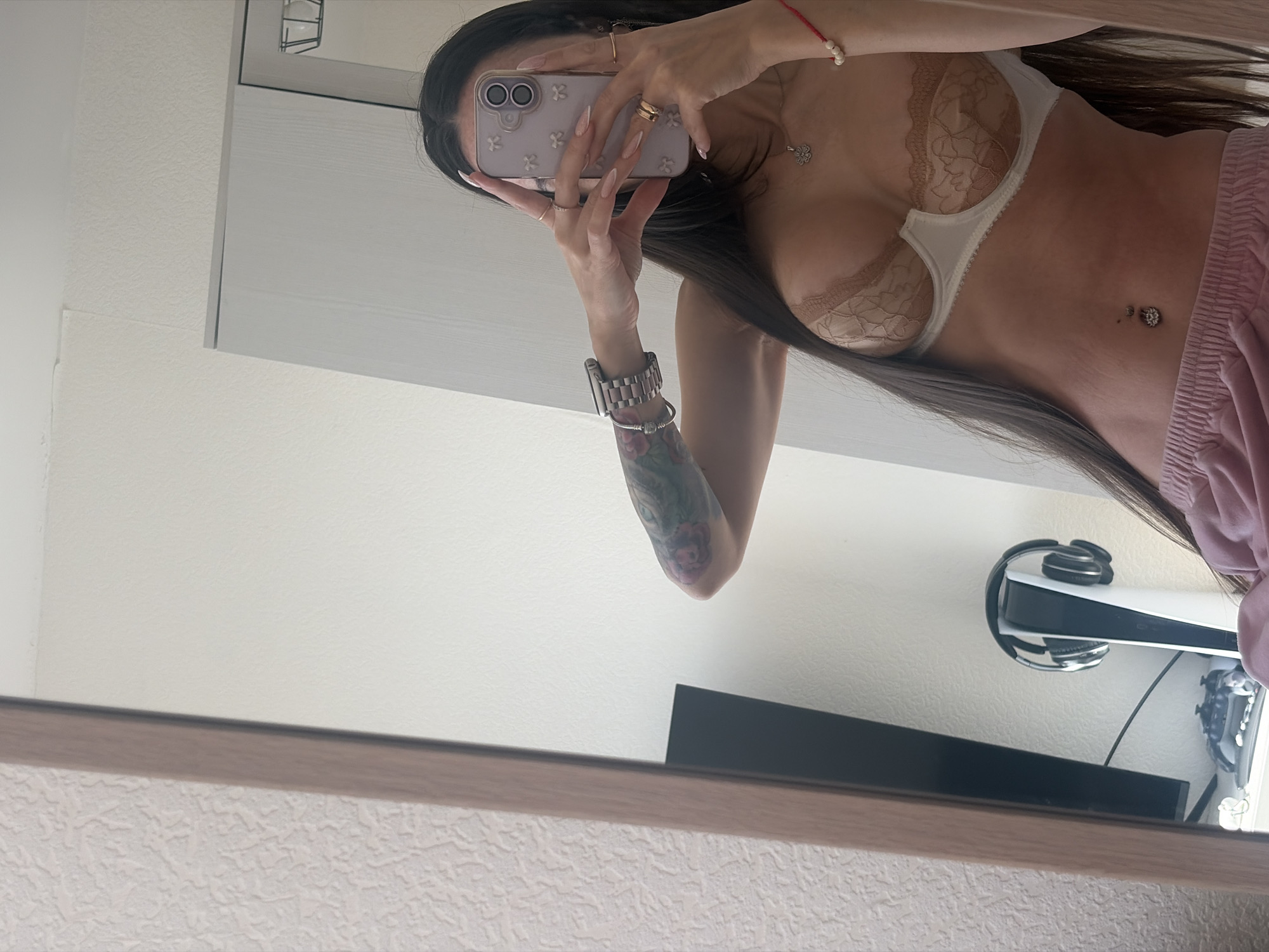 A mirrored selfie shows a woman in a bra and pink shorts. She holds a phone, obscuring her face, with a view of her upper torso. Her left arm is visible, displaying a tattoo and a wristwatch. On the wall behind her, a TV is mounted above a PlayStation 5 and headphones. The mirror's frame is in the foreground. The scene is in a room, but the location is not Saint Petersburg, Russia.