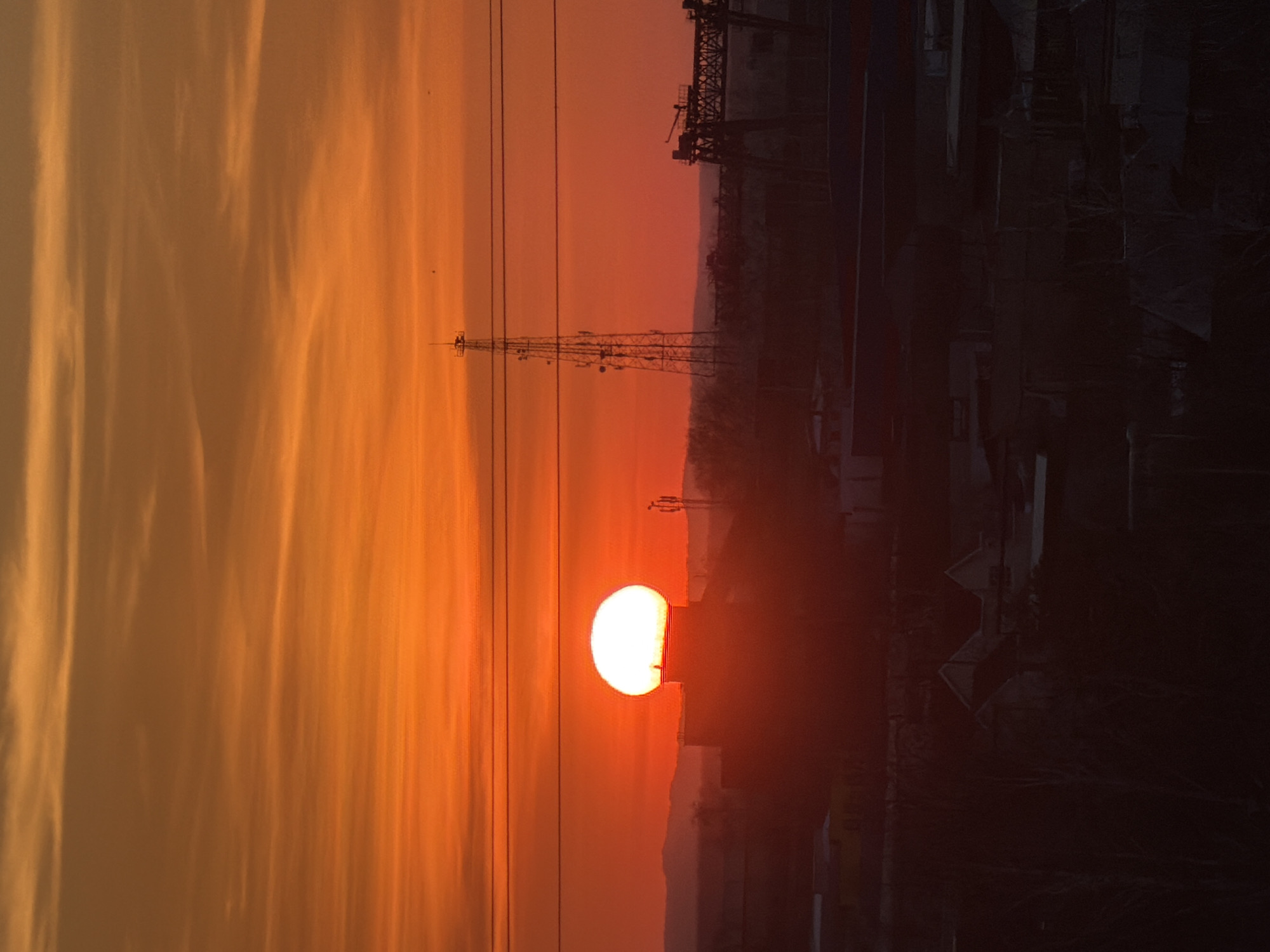 The image captures a sunset in Chaikovsky, Russia, silhouetting a radio tower and nearby industrial structures against a vibrant orange sky. Power lines stretch across the scene, adding to the industrial landscape. The sun is low on the horizon, partially obscured by the buildings, creating a bright glow. The sky displays striated cloud formations.