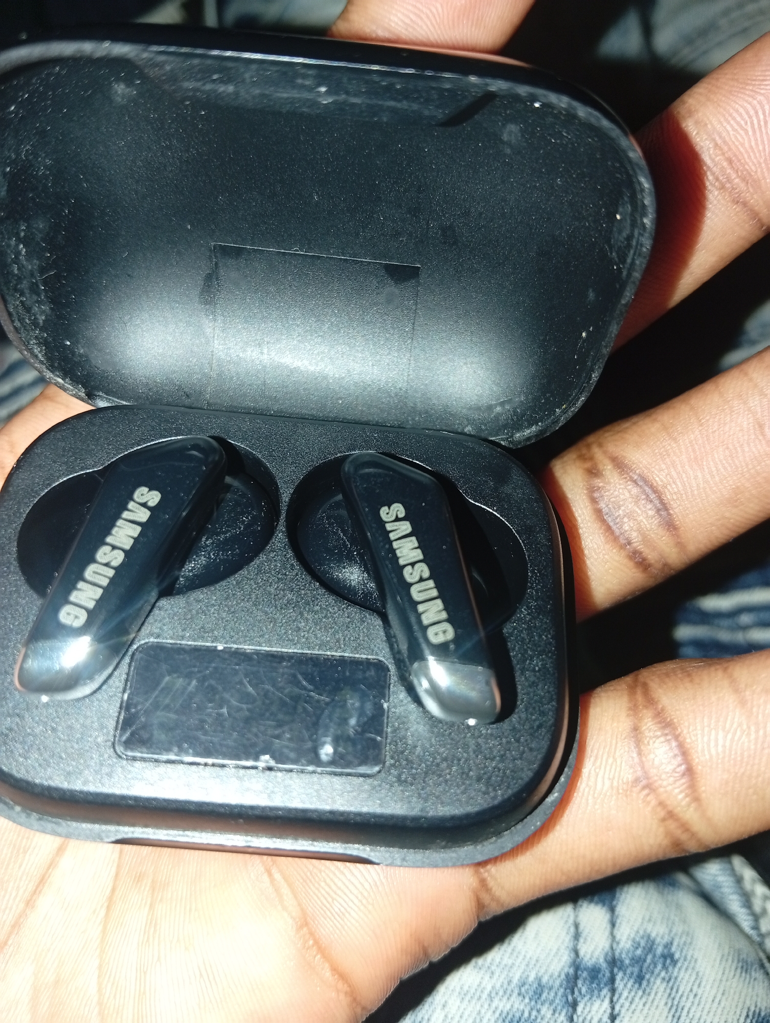 The image shows a pair of black Samsung wireless earbuds inside their charging case. The earbuds have the Samsung logo printed on them. The charging case is open, and it is being held by a person's hands, which are visible in the foreground. The background is dark and out of focus. The location in the city of Ukukwa, Nigeria is not discernible from the image itself.