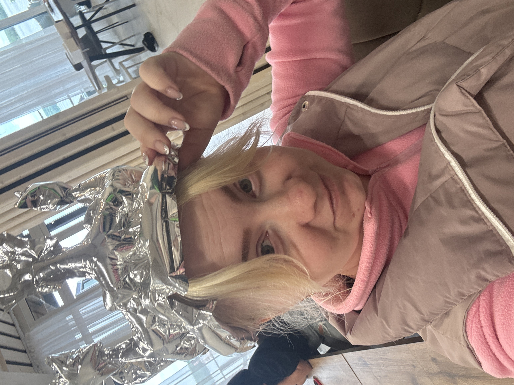 A selfie shows a blonde woman wearing a pink fleece sweatshirt and a light brown quilted vest. Her hair is covered in aluminum foil. She is indoors, near a window. Furniture is visible in the background. The location is Chaikovsky, Russia.