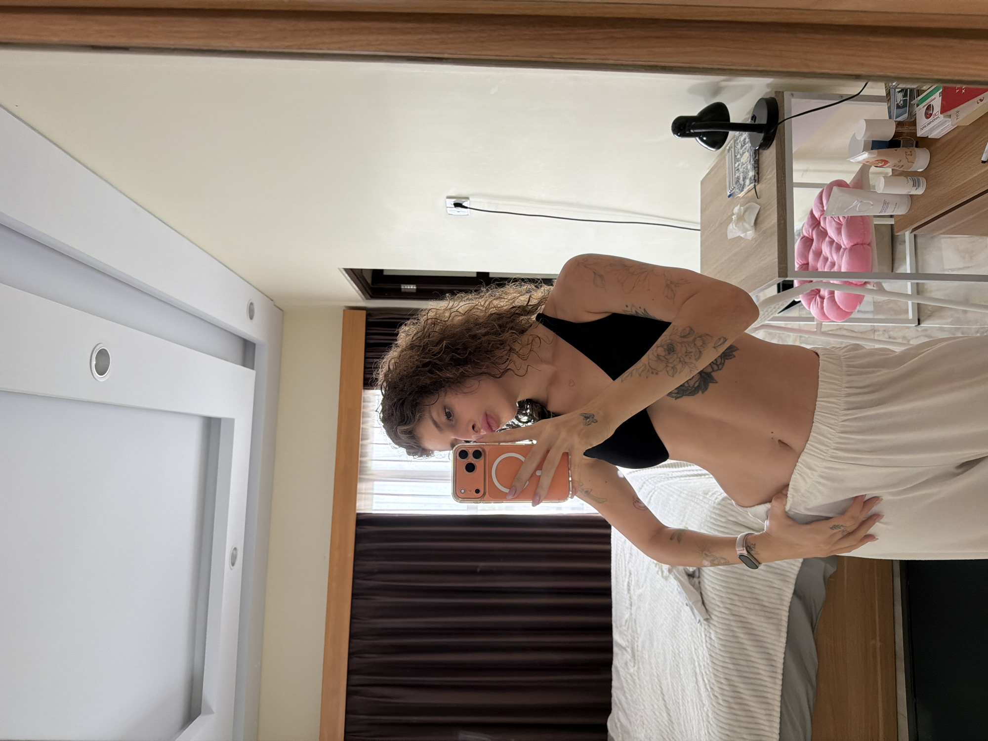 In a bedroom in North Kuta, Indonesia, a woman with curly brown hair is taking a mirror selfie. She is wearing a black bralette and white pajama pants. She has tattoos on her arms and torso. In the background, there is a bed, a desk with a lamp and some toiletries, and a window with brown curtains. The ceiling has recessed lighting.