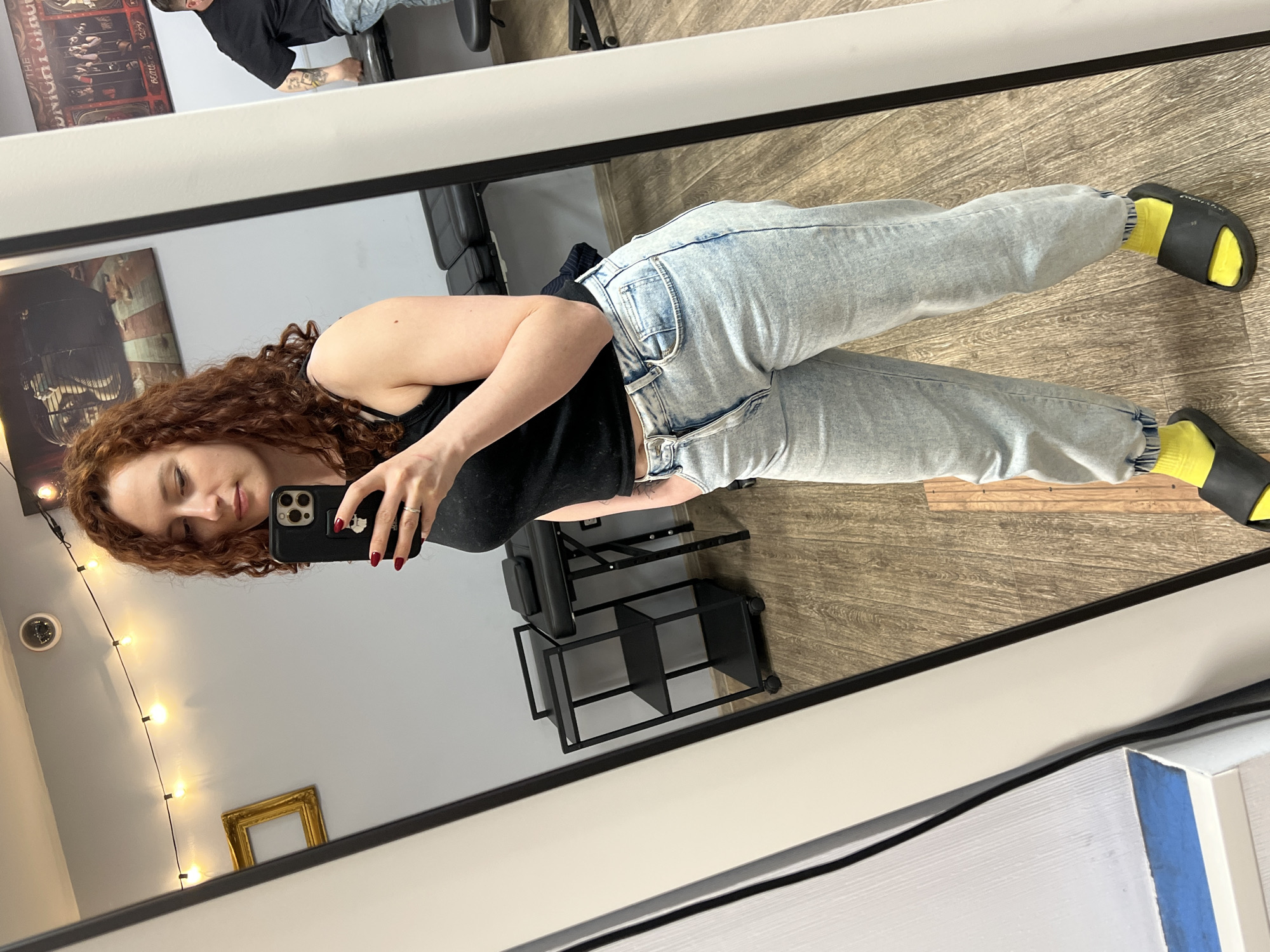 A red-haired woman in a black tank top and light blue jeans takes a mirror selfie. She is wearing yellow socks and black slides. The reflection shows a room with a black rolling chair and storage unit. The wall behind her has string lights and a gold frame. The location is likely an interior space but there is no indication it is in the city of Saint Petersburg, Russia.