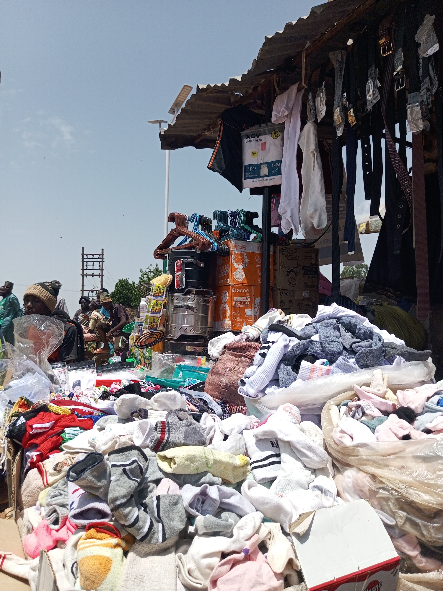 This media file depicts an outdoor market scene, likely in Bukuru, Nigeria, with a clear blue sky suggesting daytime with pleasant weather. The foreground is filled with a large assortment of clothing items, primarily socks and other small garments, piled high and overflowing from plastic bags and containers. The variety of colors and patterns indicates a bustling retail environment.
Behind the merchandise, a makeshift stall structure is visible, with a corrugated metal roof offering shade. Hanging from the stall are numerous belts, some with decorative buckles, and various pieces of clothing like white shirts. Several orange cardboard boxes are stacked, with one clearly labeled "ecoO THE COAL POT IN WEST AFRICA."
In the mid-ground and background, several individuals are present, some appearing to be vendors and others customers. They are dressed in casual attire, and some are seated, engaged in conversation or attending to their stalls. The overall atmosphere suggests a typical market day, characterized by commerce and community interaction. The visible text on the boxes and product packaging provides specific details about the goods being sold, enhancing the contextual understanding of the scene. The image captures the vibrant and everyday life of a Nigerian market.