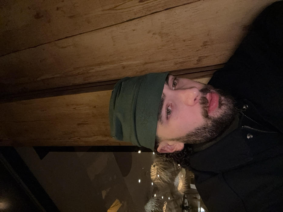 A man with a beard and green beanie is looking at the camera. He has light green eyes and fair skin. Behind him is a wooden surface and a dark, reflective area, possibly a window with lights visible. The man is wearing a dark-colored jacket with a high collar. The image is oriented sideways.