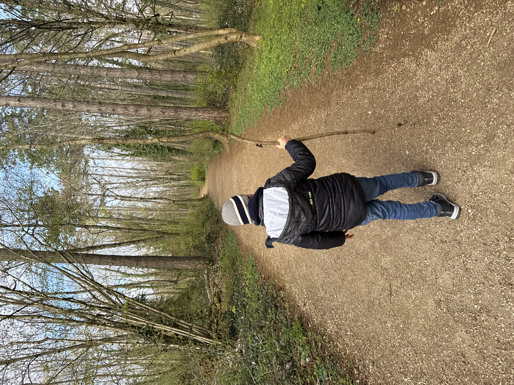 A child walks on a gravel path through a wooded area. The path stretches into the distance between tall trees on both sides. The child is wearing a black jacket, blue jeans, and a grey and white striped hat, and they are holding a long stick. The location is Esch-sur-Alzette, Luxembourg.