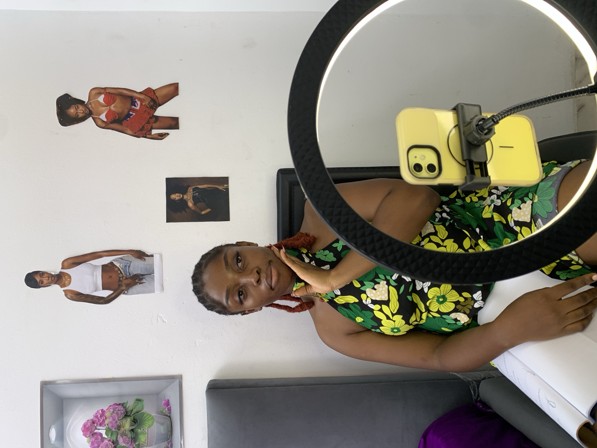The image depicts a young woman seated in front of a white wall in what appears to be a residential setting in Lagos, Nigeria. The woman, with braided hair and prominent earrings, is wearing a green and yellow floral print halter top. Her left hand is gently resting on her chin, and her right hand is visible, resting on a large open book. In the foreground, a yellow smartphone is mounted on a flexible gooseneck attached to a ring light, suggesting she might be recording or filming content. The wall behind her is adorned with several posters: a framed picture of pink flowers on the left, a cutout of a woman in a white tank top and jeans, a smaller cutout of a woman in a black dress, and another cutout of a woman in a red bikini top and a matching skirt. A gray headboard is partially visible to the left of the frame, and a purple cushion is behind it. The overall scene suggests a personal creative space, possibly a home studio, within Lagos.