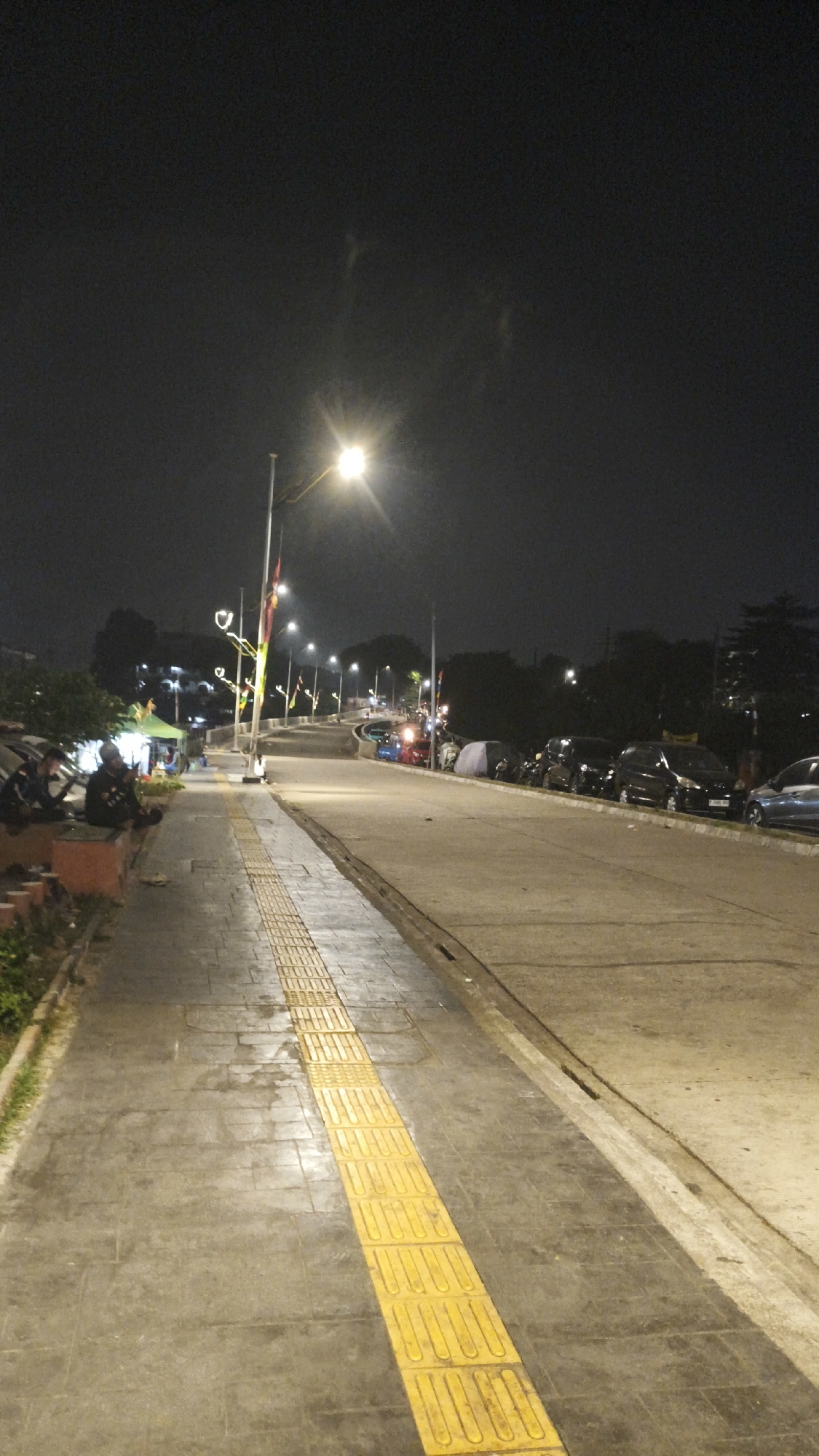 The image depicts a street scene at night in a city, possibly South Jakarta, Indonesia, illuminated by streetlights and the glow from shops or food stalls. A paved sidewalk runs along the left side of the image, featuring a prominent tactile paving strip for the visually impaired. Several people are seated on a low wall on the left side of the sidewalk, appearing to be socializing. Beyond the sidewalk, tents and temporary stalls suggest a market or event.
Across from the sidewalk, a paved road is lined with parked cars. The road curves into the distance, where more streetlights and decorative lights are visible. The overall atmosphere is one of a lively urban night, with artificial light sources creating a warm glow against the dark sky. The environment appears to be an outdoor public space, likely a street or promenade, with commercial activity and pedestrian areas.