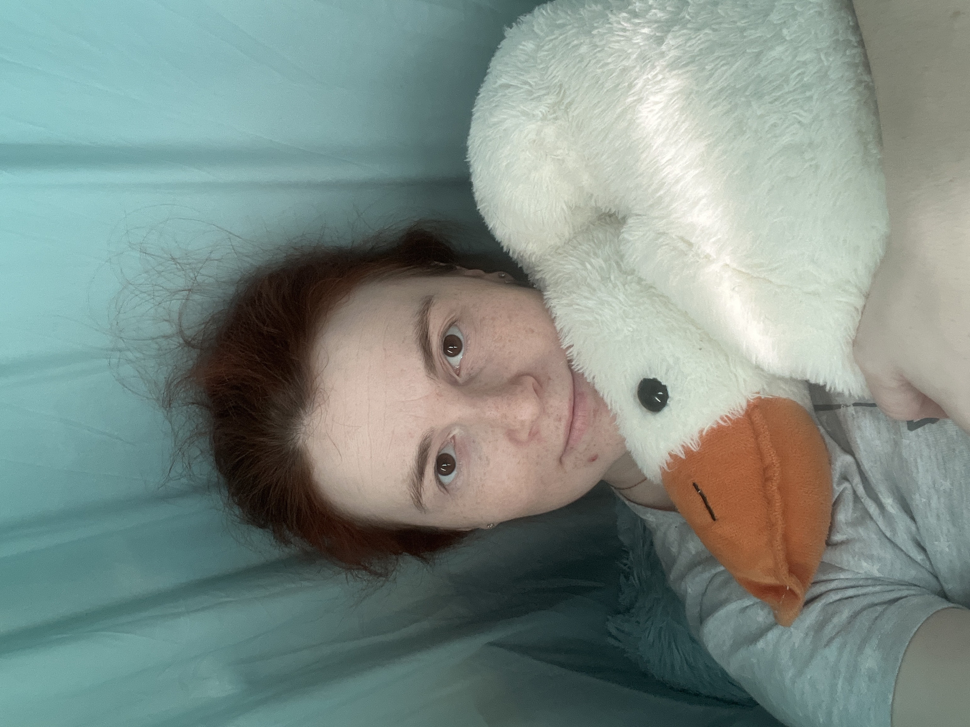 A person with reddish-brown hair and freckled skin is lying down, facing the camera. They have a plush white swan toy nestled near their face. The person is wearing a gray t-shirt. The background appears to be a tent with light blue-green fabric. The location of the photograph is not indicated in the photograph; it is not possible to determine from this image if the location is Chaikovsky, Russia.