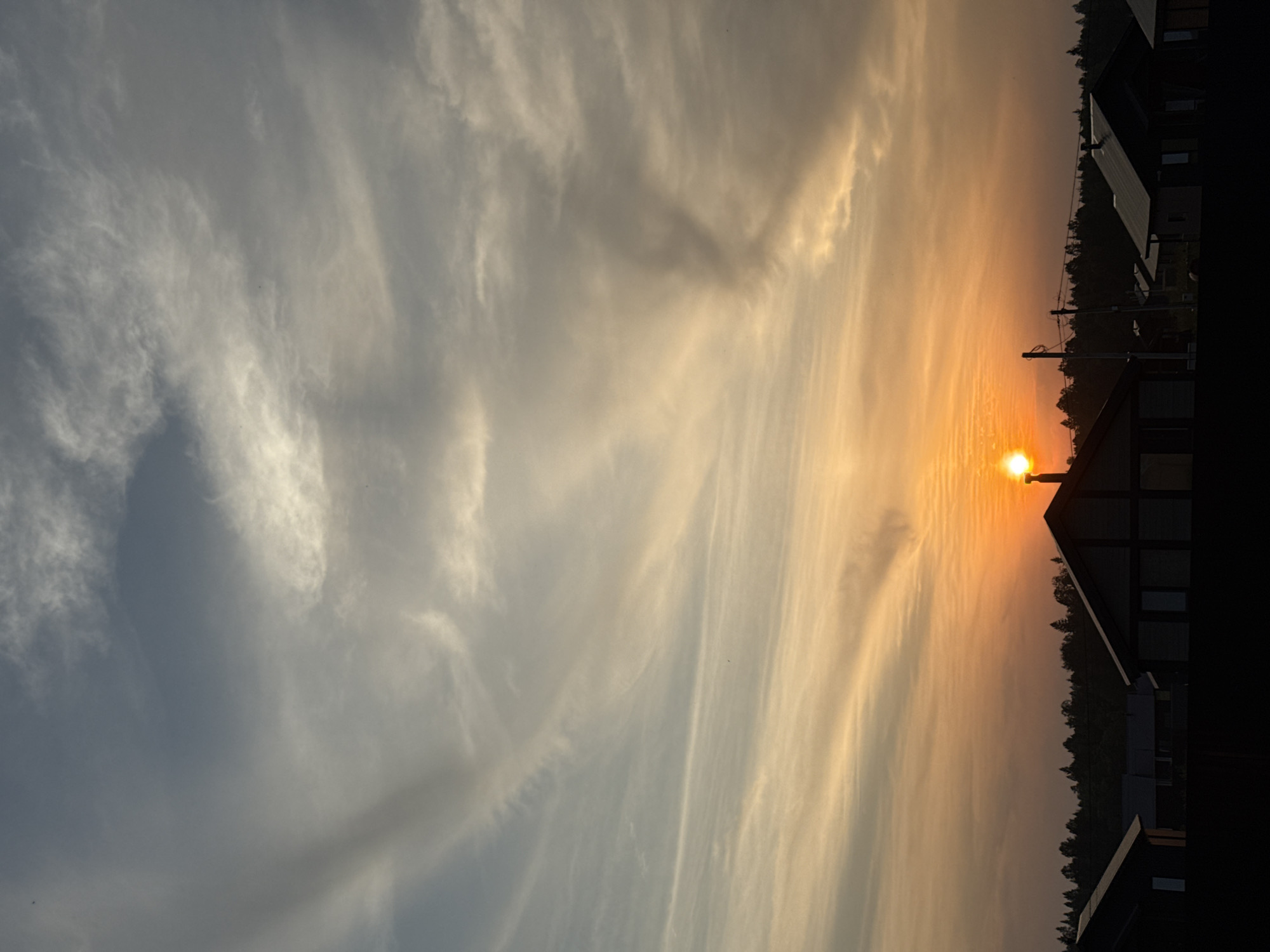 The image captures a sunset over the village of Podshivalovo, Russia. Silhouetted against the vibrant orange and yellow hues of the setting sun are several houses with dark roofs and windows. A dark line of trees forms the horizon behind the houses, with the sun appearing just above them to the left of the frame. The sky above is a canvas of wispy, striated clouds in shades of grey, white, and pale yellow, with the sun's brilliant light reflecting off some of them. The overall scene suggests a peaceful, rural evening.