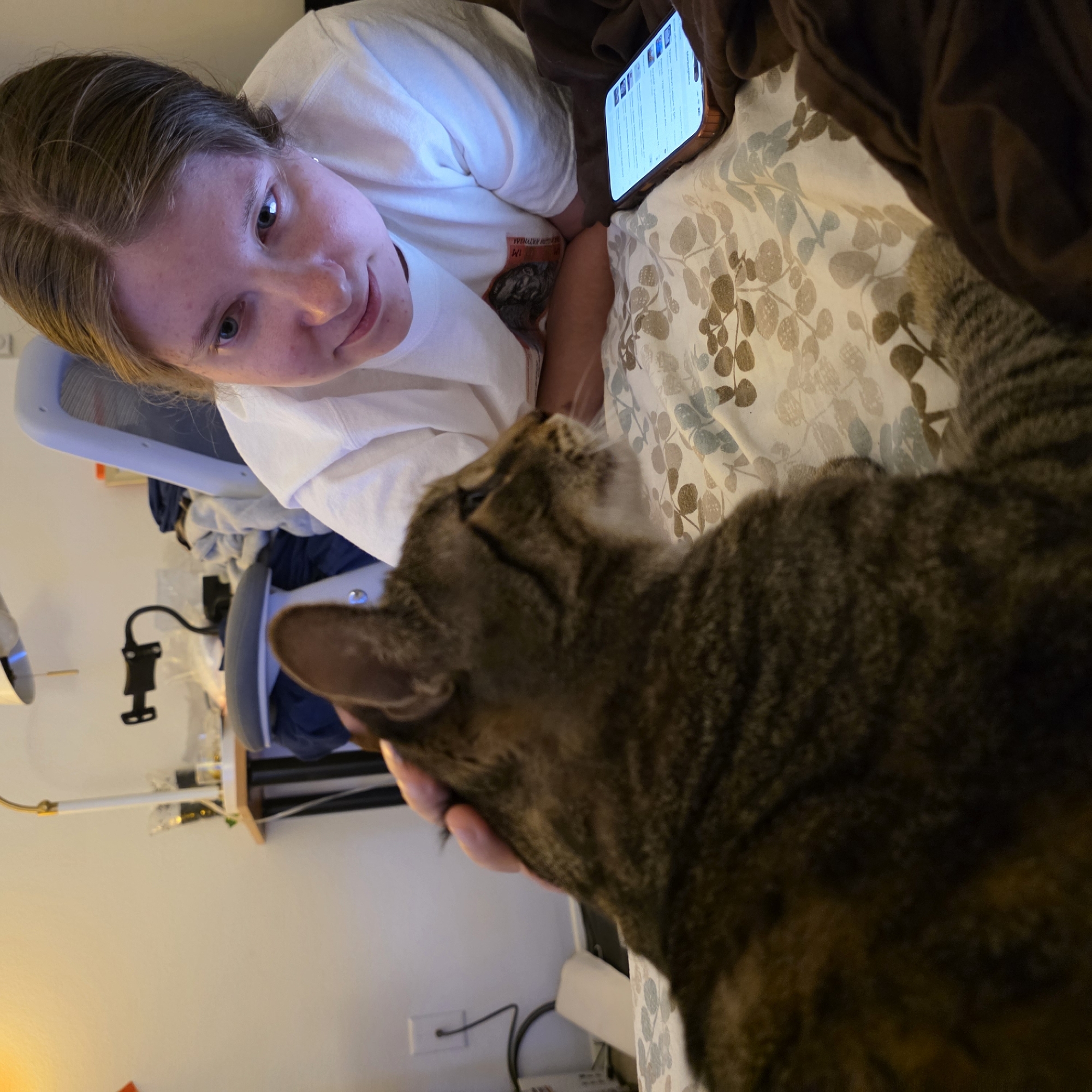 In a bedroom setting, a young woman with blonde hair, wearing a white t-shirt, is looking down at a smartphone placed on a bed. Beside her, a tabby cat is being petted by the woman's hand. The bed is covered with a patterned comforter. In the background, a desk with a lamp, a computer monitor, and some clothing is visible. The scene appears to be indoors, likely in an apartment or house. There are no external environmental cues to indicate a specific location within Portland, Oregon, or any other city.