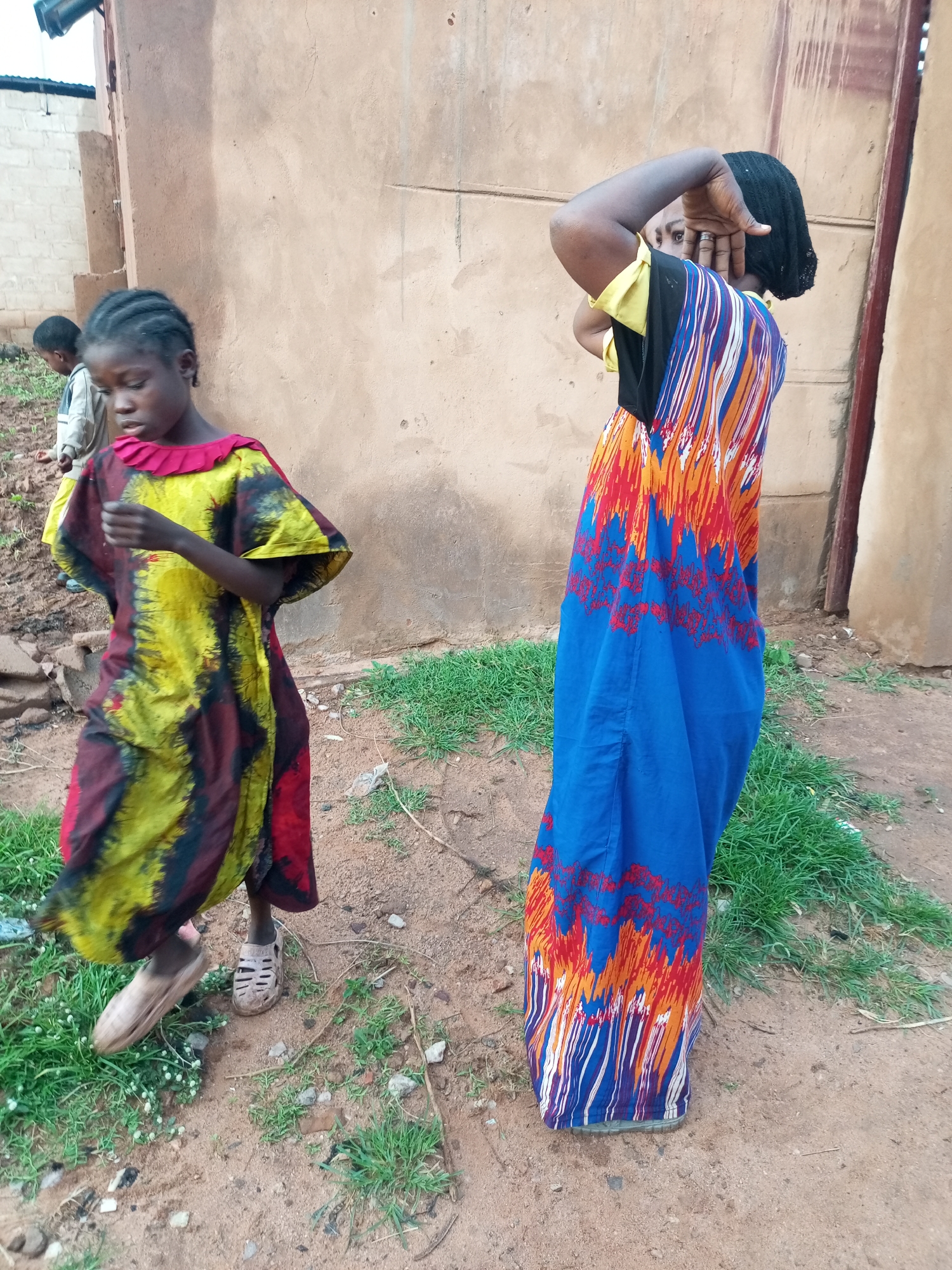 Two young females are depicted in an outdoor scene. The female in the foreground, on the left, is wearing a colorful, flowing dress with red, yellow, and black patterns. She has her hair braided and is looking downwards, with her right arm bent upwards. She is wearing light-colored sandals. To her left and slightly behind her, a second, older female is visible. She is wearing a long, blue dress with a vibrant pattern of red, orange, and white lines. She has a black headscarf and is shielding her face with her right hand. Both females are standing on a dirt ground with sparse green grass. In the background, a wall of a building with a rough, textured surface is present. To the far left, a portion of another building with white brickwork is visible. The overall setting appears to be an urban environment in Tempure, Nigeria.