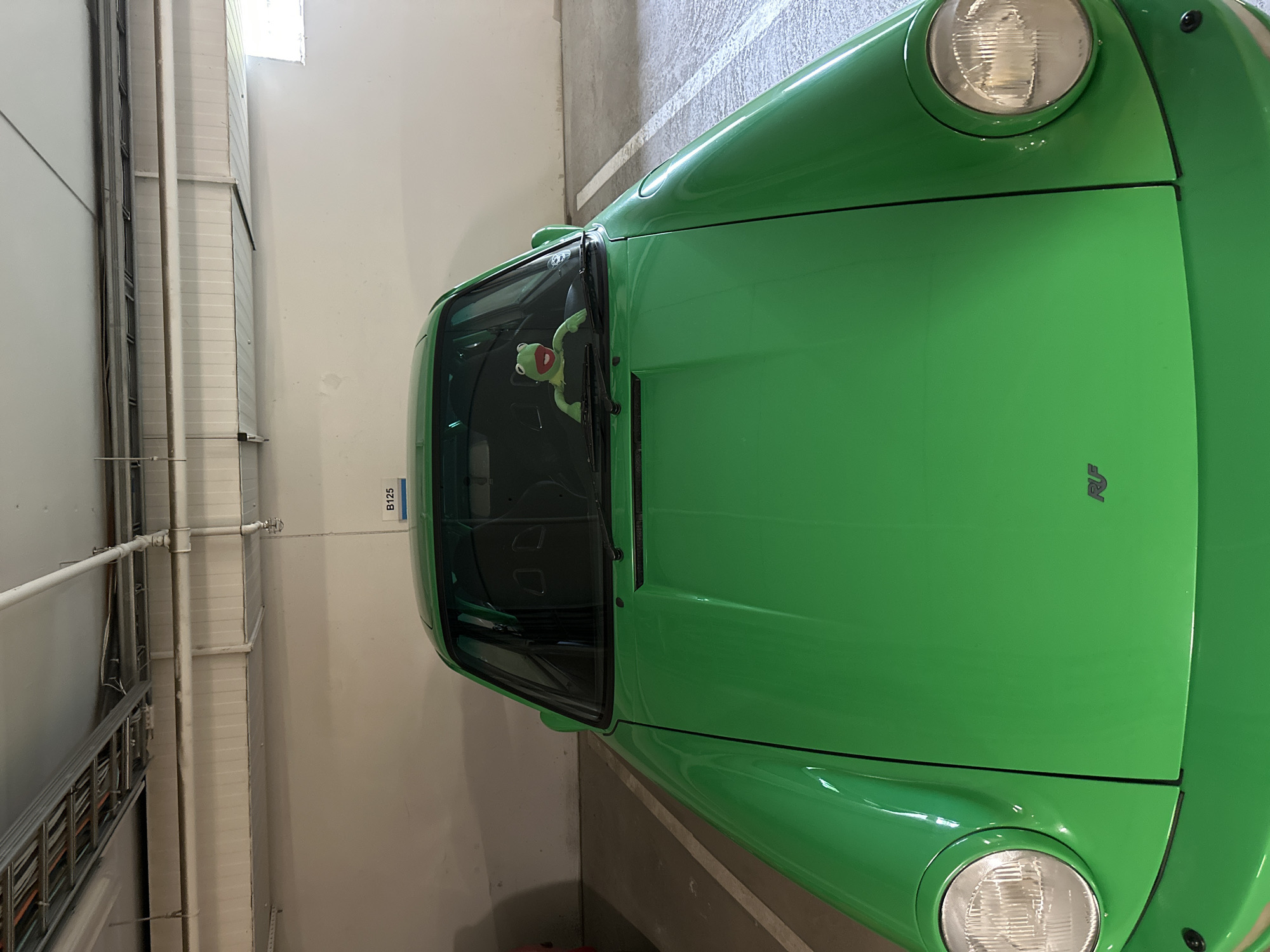 A Kermit the Frog plush toy is positioned on the dashboard of a bright green car, likely a Porsche 911 based on its design, parked in an indoor parking garage. The car is centrally framed and occupies most of the image. The parking garage features white walls, a concrete floor with white parking lines, and an overhead structure with pipes and lighting fixtures. A sign labeled "B125" is visible on the wall behind the car. The overall lighting suggests an indoor, artificial environment. The scene is static, with no visible activity or interaction. This image was taken in Espoo, Finland.