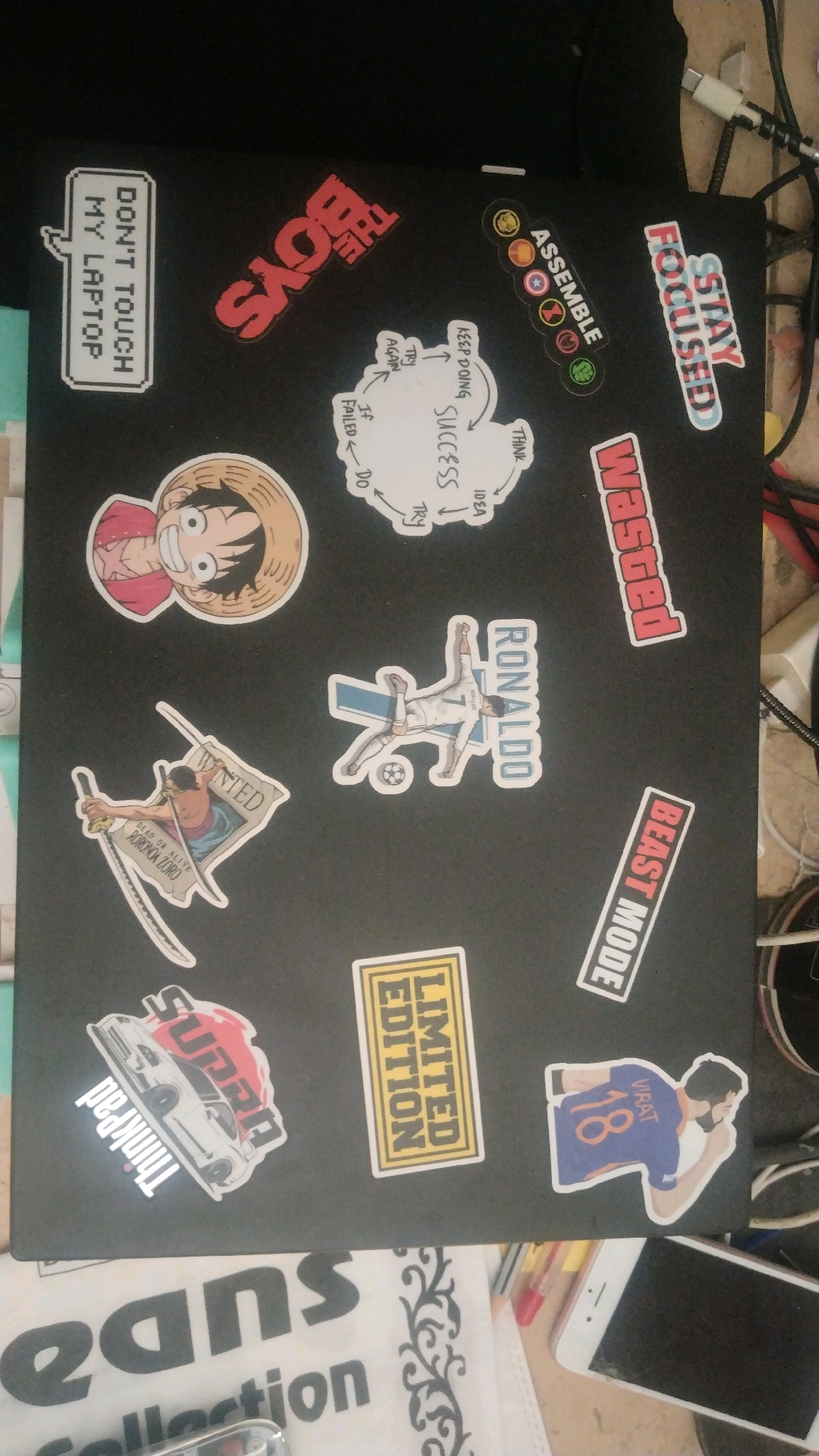The image shows the lid of a black laptop adorned with various stickers. These stickers include a portrait of Monkey D. Luffy from One Piece, a "WANTED" poster featuring Roronoa Zoro, a sticker of Cristiano Ronaldo in action, and a sticker of a Toyota Supra. There are also motivational stickers with phrases like "DON'T TOUCH MY LAPTOP," "THE BOYS," "STAY FOCUSED," "ASSEMBLE," "WEB GEEKS," "BEAST MODE," "LIMITED EDITION," and "Thinkpad." Additionally, there's a diagram illustrating a process of success. The laptop is placed on a surface that appears to be a table, with some cables and a smartphone visible in the background. The lighting suggests an indoor environment. The image is taken from a slightly elevated angle, looking down at the laptop. There is no discernible information in the image that would indicate a specific location within Bihar Sharif, India.