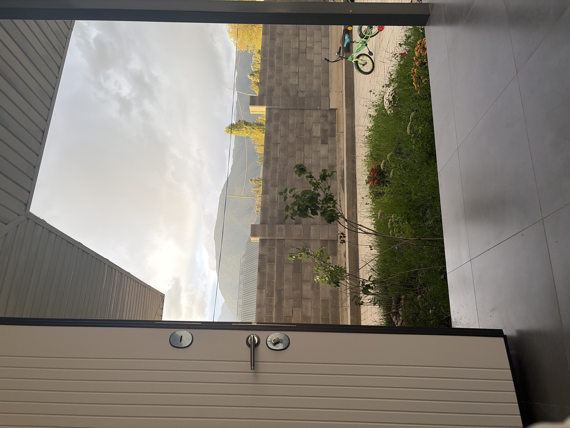 The image captures a view from an open doorway, looking out onto a street scene in Bayet, Kyrgyzstan. In the foreground, a light-colored, modern door with a silver handle and two circular lock mechanisms is partially open on the left. The floor in front of the door is tiled with large, square, gray tiles. Beyond the doorway, a green lawn with scattered wildflowers is visible. A grey concrete block wall runs horizontally across the mid-ground, separating the immediate area from the street beyond. On the other side of the wall, a street is visible, and further back, a small green bicycle with a yellow seat and red accents is parked. Trees with yellow and green foliage line the street, and in the distance, rolling hills or mountains under a cloudy sky dominate the horizon. The overall impression is of a residential area on a cloudy day.