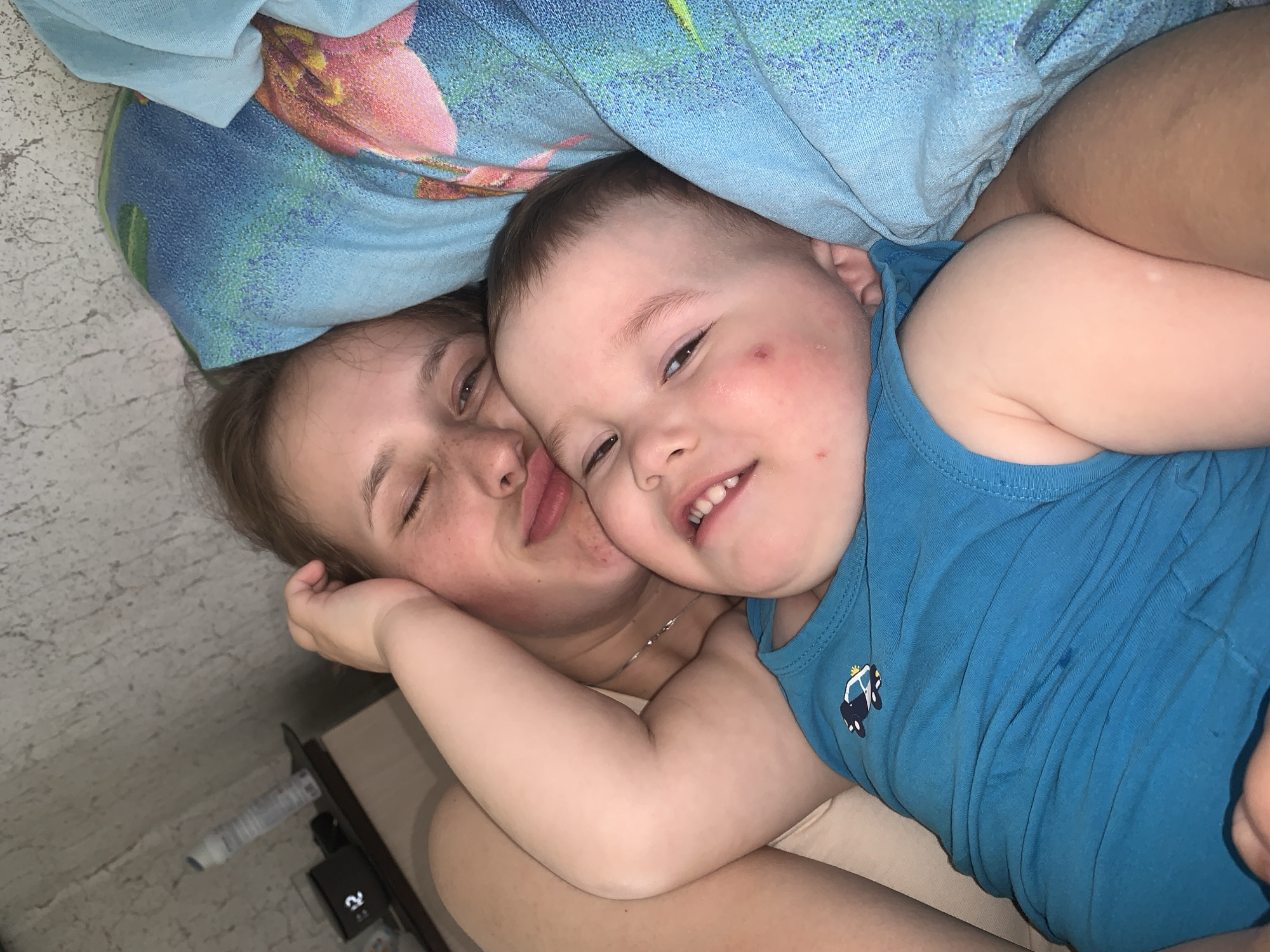 A photograph shows a person with brown hair and closed eyes and a child with a police car on their blue shirt lying down next to each other under a blue and pink floral blanket. The location in the city of Фоки, Russia is not identifiable in the image.