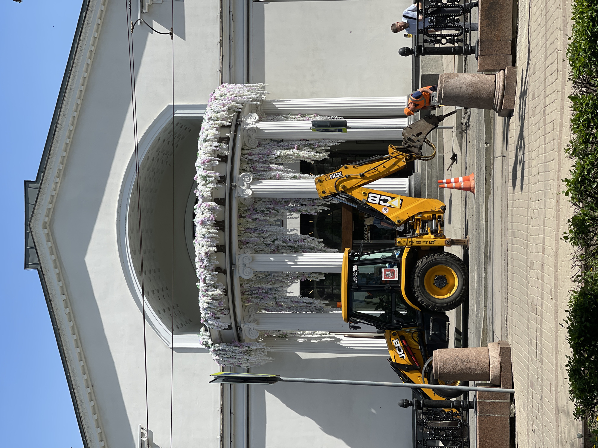 The image depicts a JCB backhoe parked on a city street in front of a building, which is decorated with white and lavender flowers and has white columns. A construction worker in a hard hat is using a jackhammer near the JCB. A man is standing on a balcony overlooking the scene. The street is paved with brick. Trees are visible on the right side of the frame. The city is Unknown, Unknown.