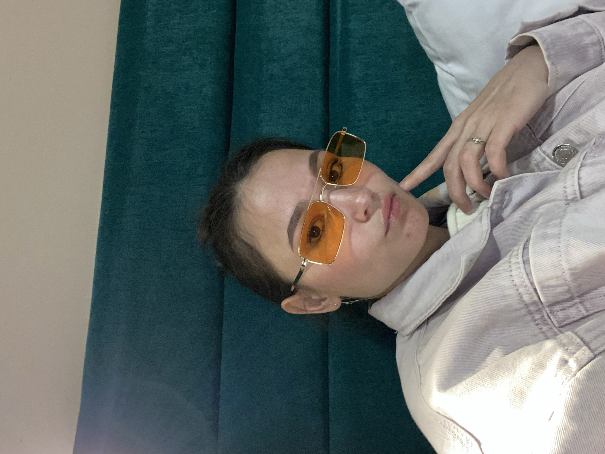 A woman is lying down on a surface near a turquoise curtain. She is wearing orange tinted glasses and a light purple denim jacket. Her left hand is near her face, showing a ring on her finger. The background appears to be a wall, partially covered by the curtain. The location of Peresvet, Russia cannot be determined from the image.