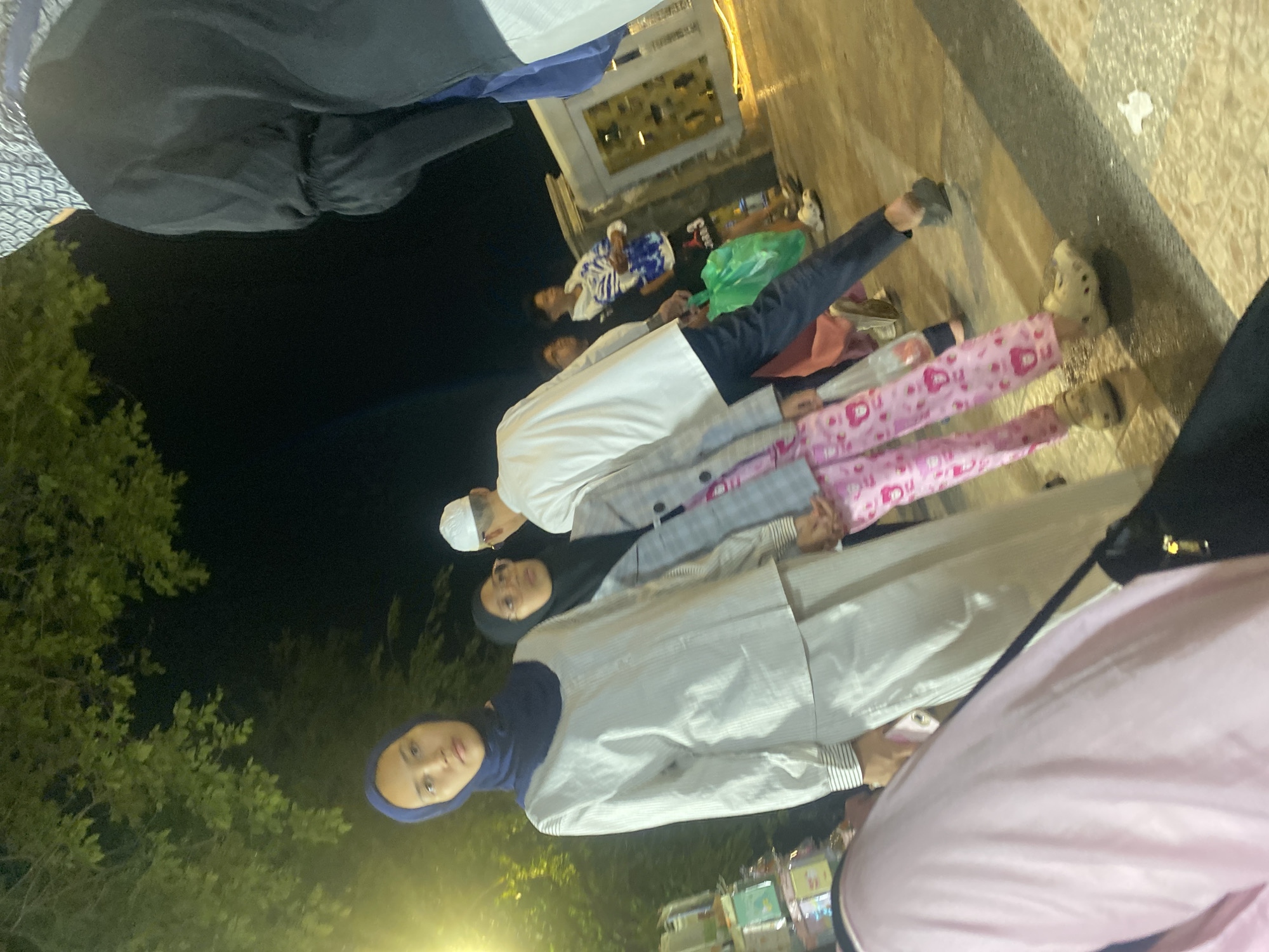 In Banjarmasin, Indonesia, at night, several individuals are gathered outdoors. In the foreground, a woman wearing a dark blue hijab and a light-colored striped outfit stands facing forward, with a black bag on her left side. To her right and slightly behind her, a younger person in a black hijab and a checkered grey and white blazer over pink patterned pants, also in a hijab, is visible. Another individual, possibly an adult male, wearing a white long-sleeved shirt and a white cap, stands further in the background and to the right of the two foreground individuals, facing away. Further to the right, other individuals are present, including a person holding a bright green plastic bag. The ground appears wet, reflecting the ambient light. Trees with green foliage are visible in the upper left portion of the image, against a dark night sky. Various items are stacked to the left of the foreground subjects, possibly merchandise on display.