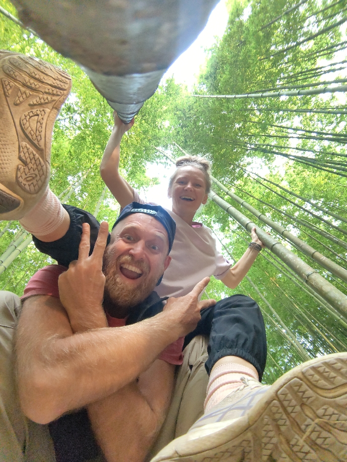 An extreme low-angle, wide-perspective photograph depicts a smiling Caucasian man supporting a Caucasian woman in a dense bamboo forest in Mù Cang Chải, Vietnam. The man, positioned in the lower center, has a light brown beard, wears a dark baseball cap backward, and a reddish-pink t-shirt. He displays a wide, open-mouthed smile and makes a "peace" sign with both hands. The woman is perched above him, partially obscured, with her light-colored athletic shoe and pink-socked foot prominent in the lower left. She wears a light pink top and black cuffed pants, has her hair in a bun, and holds onto a thick bamboo pole with her right hand, extending diagonally across the upper frame. She also exhibits a wide, open-mouthed smile. The background is filled with numerous tall, thin green bamboo stalks reaching upwards, with bright sunlight filtering through the canopy, illuminating the scene.