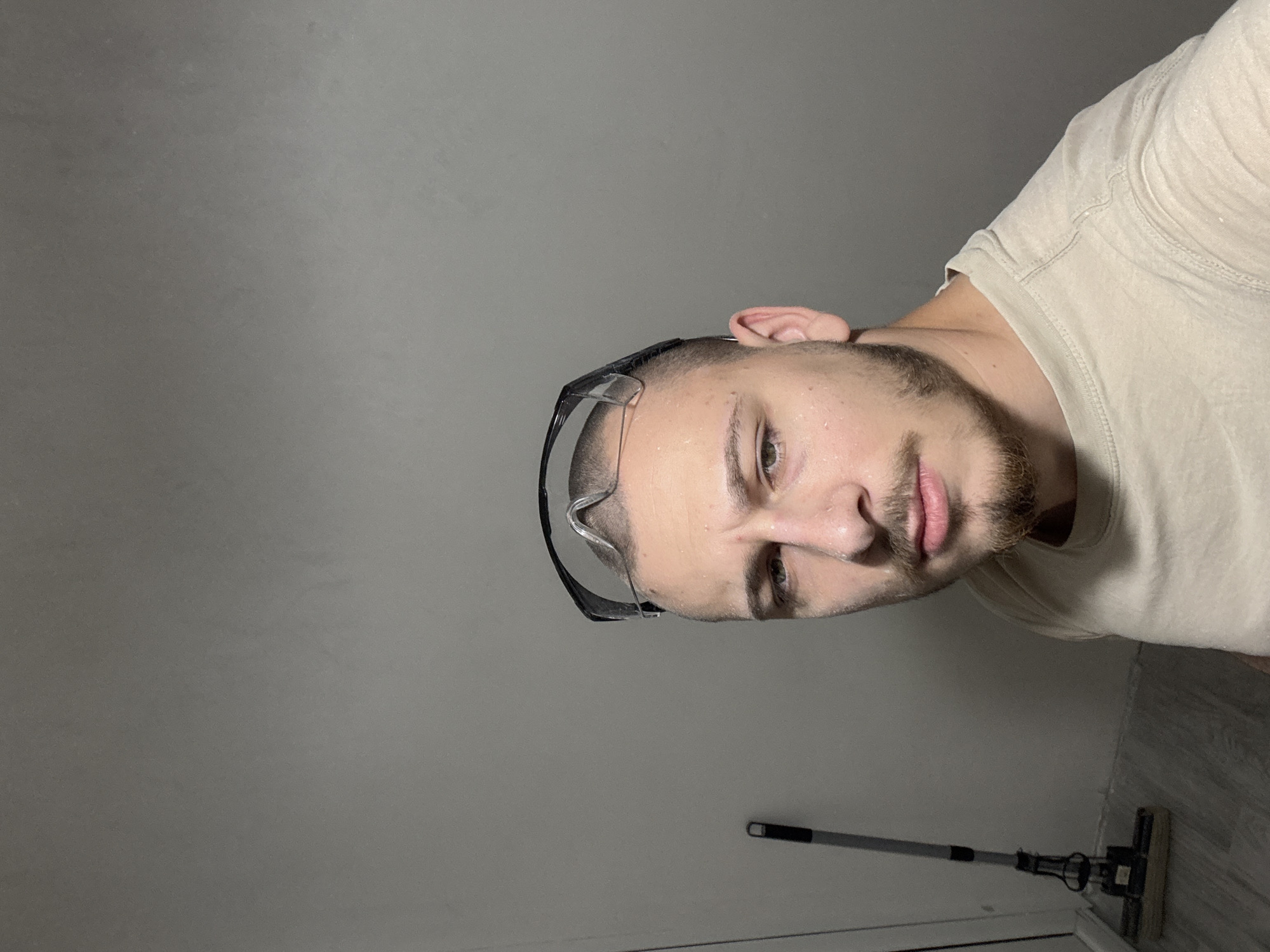 A man wearing safety glasses and a beige shirt is visible in a close-up, selfie-style shot. He has short hair, a beard, and appears slightly sweaty. The background consists of a gray wall and a glimpse of gray wood-look flooring. A mop with a gray handle is leaning against the wall. The location is not identifiable as Stara Zagora, Bulgaria based on the visual elements in the photograph.