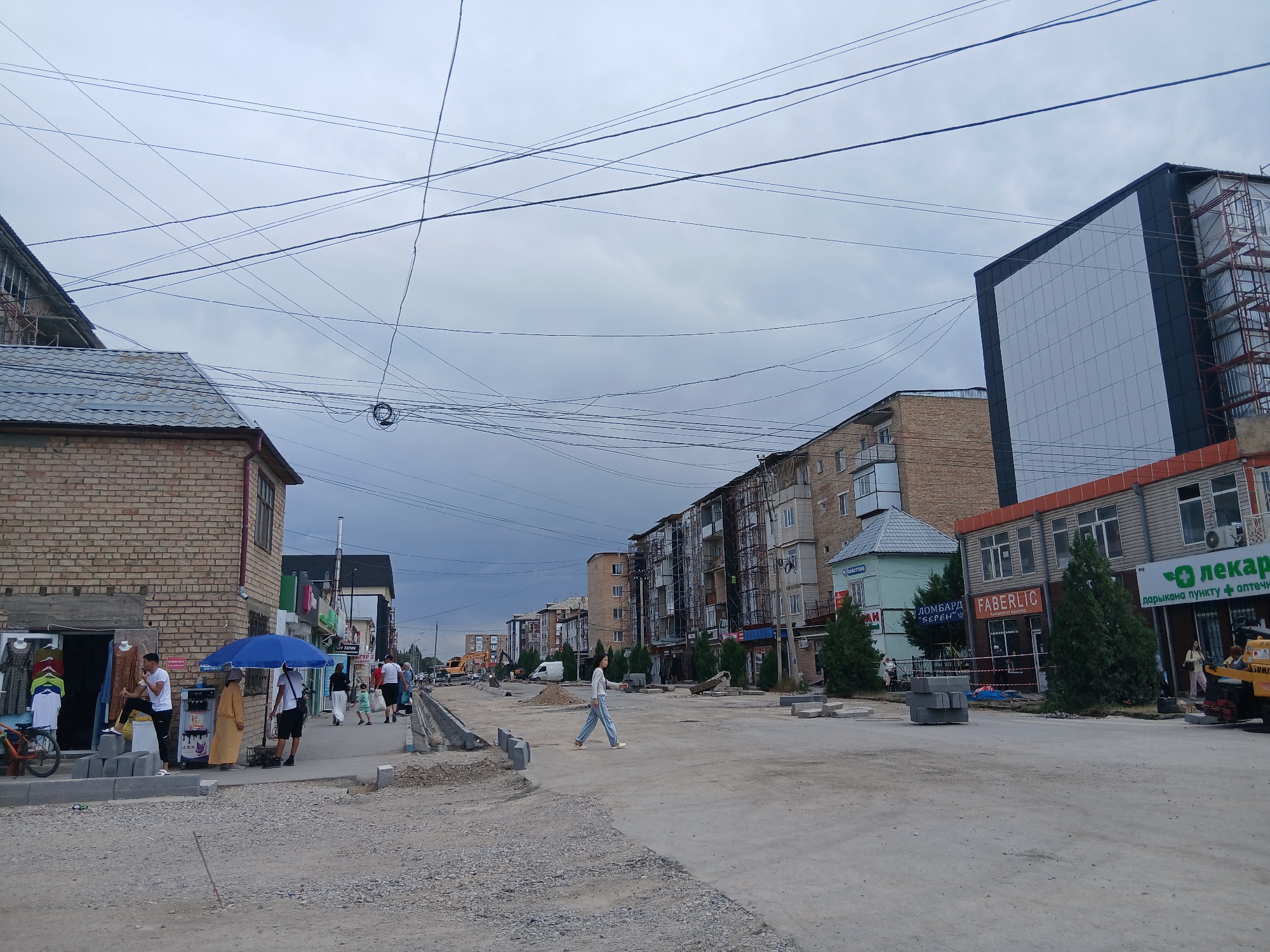 The image depicts a street scene in Talas City, Kyrgyzstan, under an overcast sky. The foreground is a dirt and gravel road, indicative of ongoing construction or roadwork, with concrete barriers and piles of materials visible. A woman in light blue pants and a white long-sleeved shirt is walking across the road from right to left.
Along the right side of the street, there are several buildings. The closest building has a modern facade with large windows and signage that includes "Faberlic" and what appears to be a pharmacy with a green cross logo. Adjacent to it, a green building with a pointed roof is visible. Further down the street, a larger building with a dark facade and many windows is under construction, with scaffolding attached.
On the left side of the street, a two-story brick building with a slanted grey roof houses a small shop. Several people are standing in front of this shop, including a man in a white t-shirt and dark pants, and another person wearing a yellow robe. A blue umbrella is open near the shop. A bicycle is parked to the left of the shop. Further down this side of the street, there are more buildings and what appears to be a construction site with heavy machinery. Power lines crisscross the sky above the entire scene.