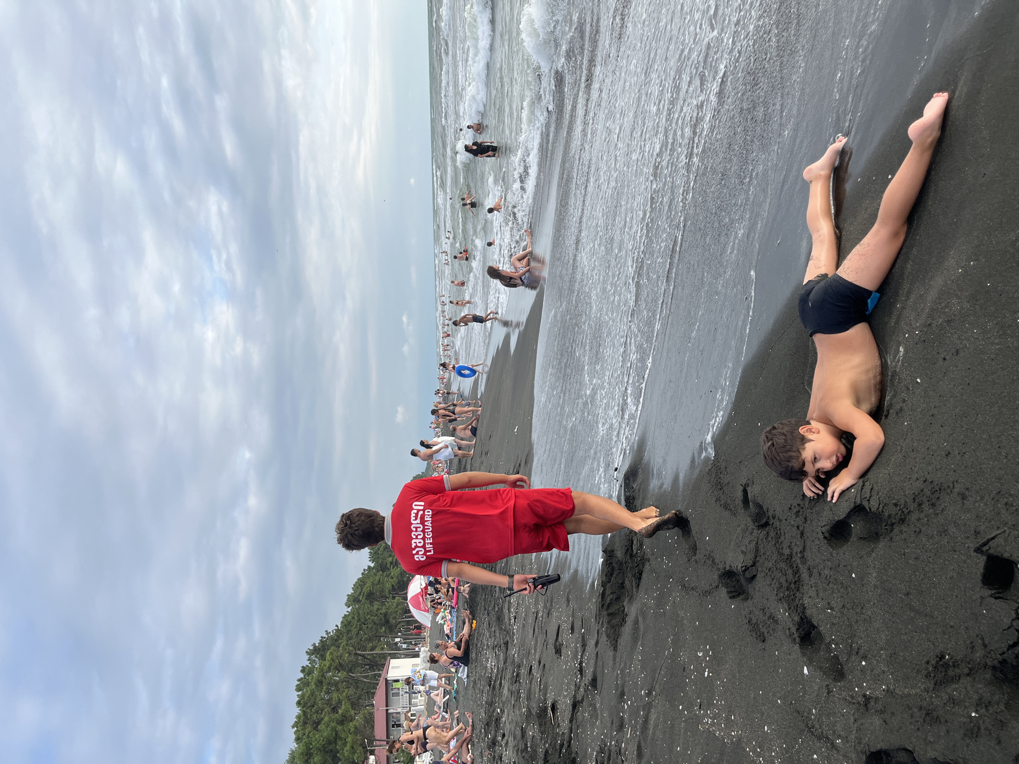 The image captures a scene at the black sand beach in Ureki, Georgia. In the foreground, a young boy lies face down on the wet sand near the water's edge, wearing black swim trunks. Behind him, a lifeguard, dressed in a red shirt with "LIFEGUARD" printed on the back and red shorts, walks away from the camera, holding a black walkie-talkie. His feet are covered in dark sand. The beach is populated with many people sunbathing on the sand, swimming in the wavy sea, or wading in the water. The sky is overcast with light clouds. The background shows a line of pine trees and some buildings.