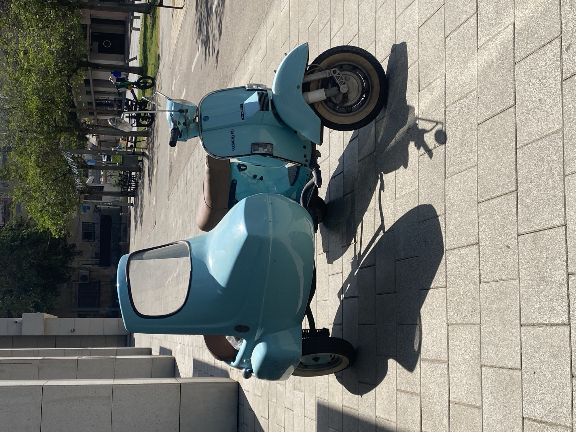 A vintage-style light blue Vespa scooter with a matching sidecar is parked on a paved urban sidewalk. The scooter features a classic design with a brown seat, a light blue body, and whitewall tires on its main wheel. The brand names "Vespa" and "Piaggio" are visible in dark lettering on the scooter's leg shield. The sidecar, also light blue, has a small, clear windshield framed in black.
The setting is an outdoor area under bright, clear skies. Strong, distinct shadows from the scooter and nearby trees are cast across the light grey, tiled pavement, indicating a sunny day, likely during mid-morning or late afternoon. To the left, a row of green trees lines what appears to be a street or pedestrian path. In the background, urban buildings are visible, some with windows and an air conditioning unit. A parked bicycle can be faintly seen near the trees. No people are visible in the image, suggesting the area might be quiet. The scene conveys a relaxed, sunny urban atmosphere, consistent with Tel-Aviv, Israel.