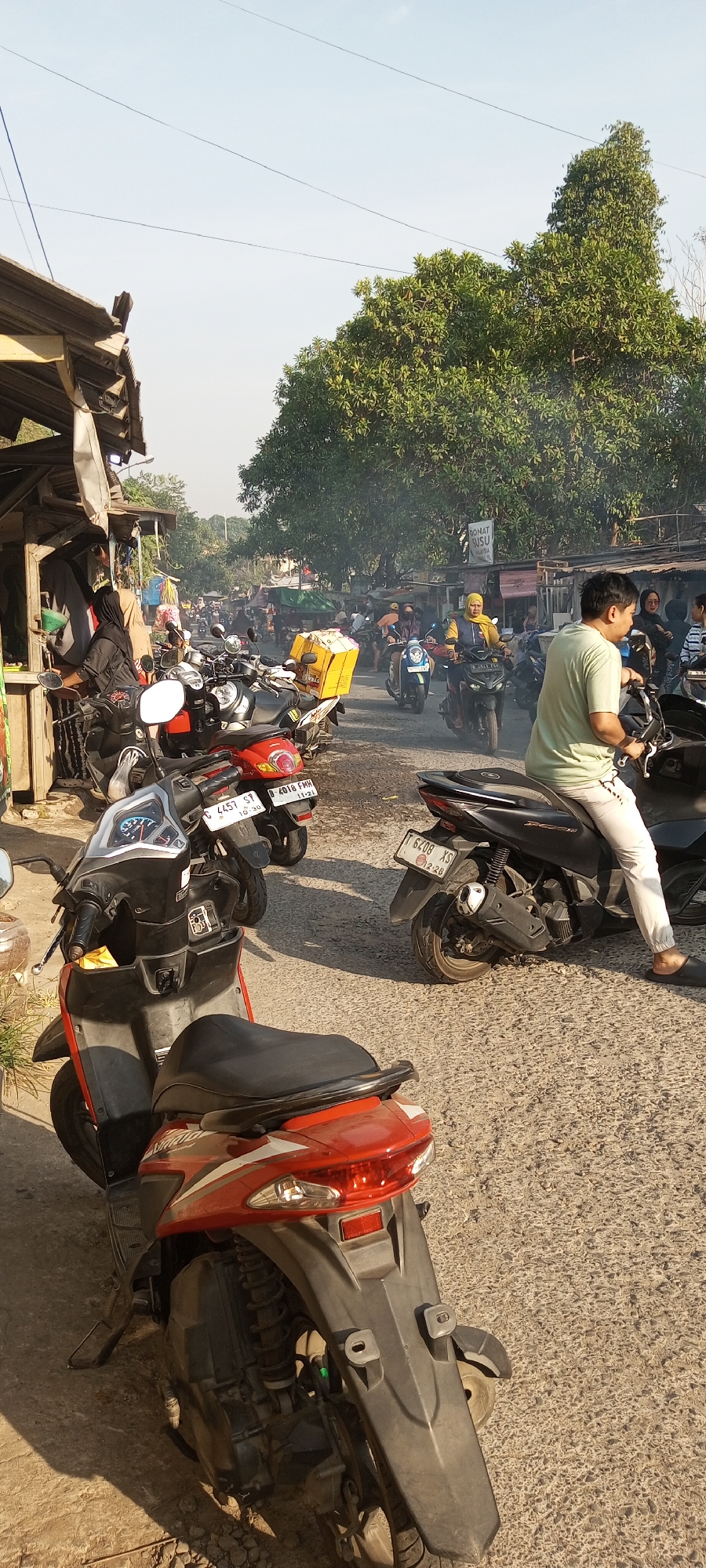 The image depicts a busy street scene in Setu, Indonesia, characterized by a significant number of motorcycles. In the foreground, several parked motorcycles are visible, one prominently red and white. A man wearing a light green t-shirt and white pants is seated on a black motorcycle, facing away from the camera.
The background reveals a bustling street with more motorcycles in motion and parked. A person wearing a yellow headscarf is riding a dark motorcycle. Buildings with makeshift stalls line the street, suggesting a market or commercial area. Yellow boxes are visible on the back of some motorcycles, possibly indicating delivery services. Smoke or dust is present in the air, creating a hazy atmosphere. The overall environment appears to be an outdoor urban setting with a mix of commercial activity and traffic. The sky is clear with some power lines crisscrossing overhead.