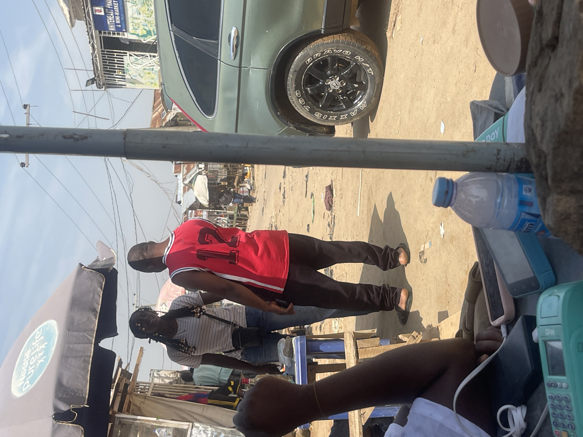 The image depicts a street scene in Abuja, Nigeria. In the foreground, a hand is raised, and a collection of mobile phones and a green electronic device are spread out on a surface. Behind this, two individuals are conversing. One person wears a red jersey with the number "12" on it, and the other wears a striped t-shirt and jeans. They are standing near a blue table. Further in the background, a green SUV is parked. The environment appears to be a marketplace or a busy street, with various stalls and buildings visible, including a sign for "WHITEHEART PHARMACY & MINI-MARKET". The ground is unpaved, and the overall setting suggests a sunny, outdoor environment.