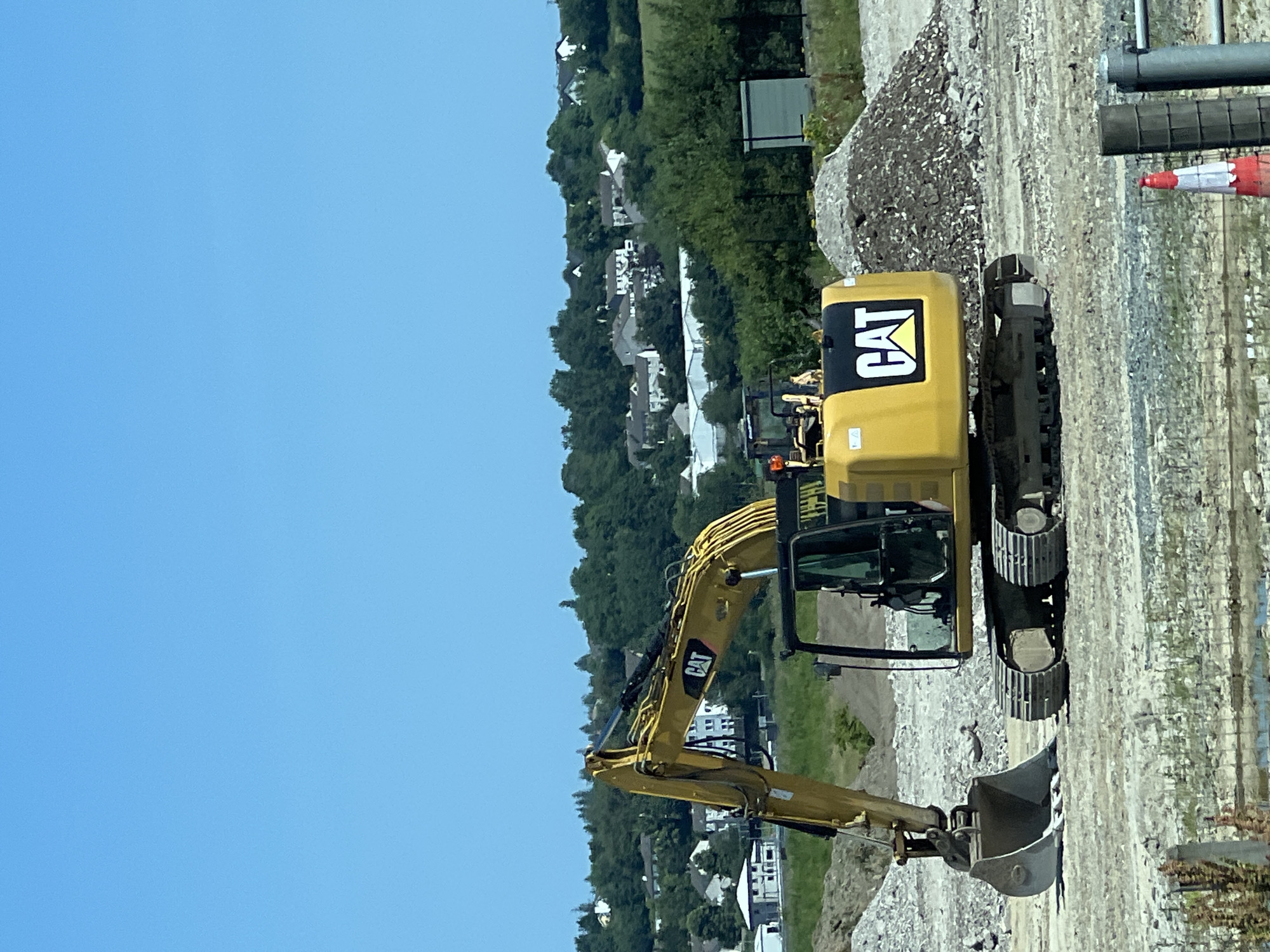 This is a photograph of a yellow Caterpillar excavator at a construction site under a clear blue sky. The excavator is positioned in the foreground, with its arm extended and bucket resting on the ground. The machine has the "CAT" logo prominently displayed on its rear. In the background, there is a landscape with rolling green hills dotted with houses, suggesting a suburban or rural setting. The ground at the construction site is a mixture of dirt, gravel, and debris. There are also piles of rubble and a large green shed visible. The overall scene depicts an active construction zone with a focus on the heavy machinery. The location is identified as Letterkenny, Ireland.