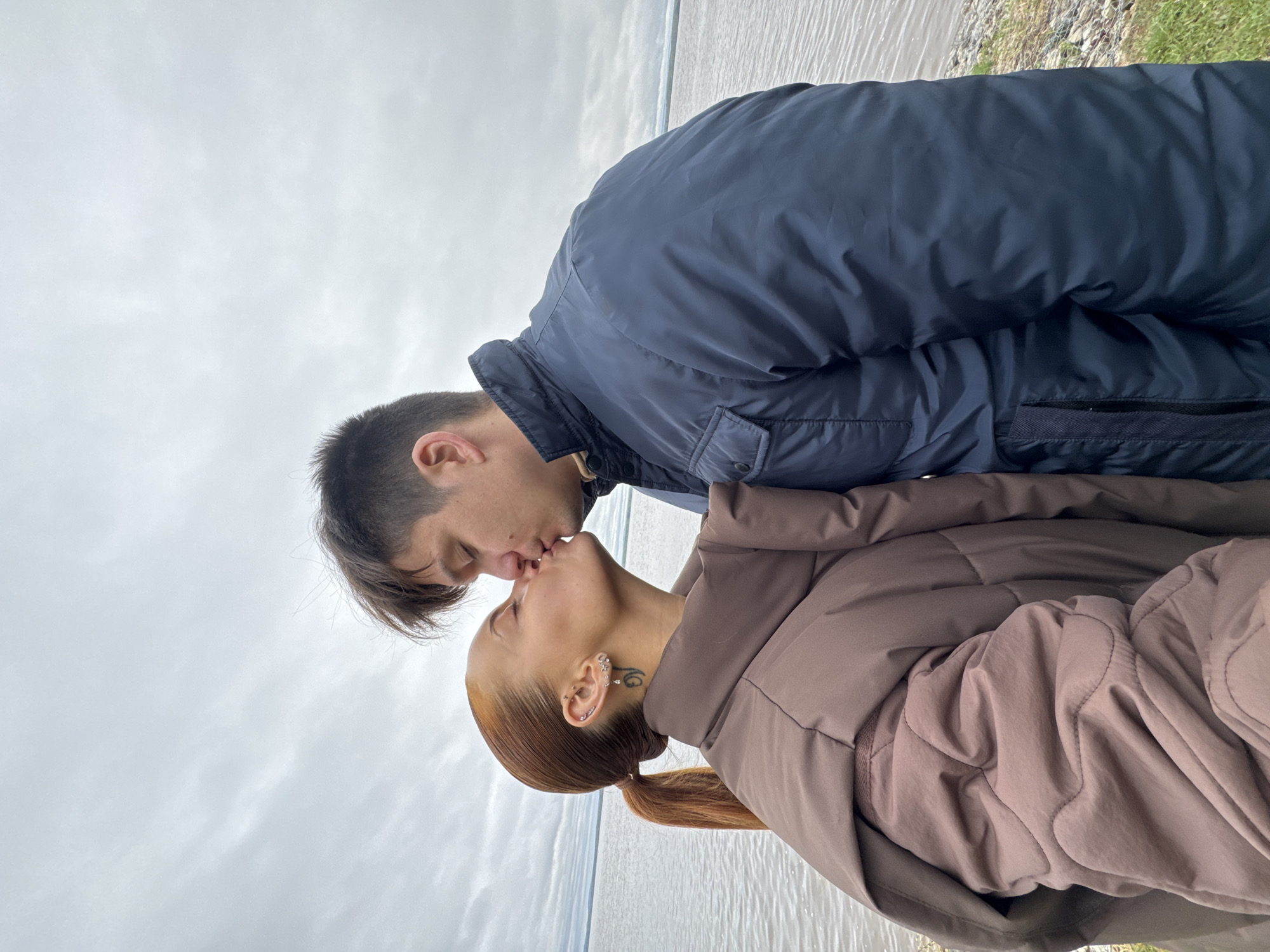 A photograph shows a couple kissing near a body of water, possibly the Kama River in Chastyye, Russia, given the landscape. The man on the left has short, dark hair and is wearing a dark blue jacket. The woman on the right has red hair pulled back into a low ponytail, a tattoo on her neck, and is wearing a brown puffy jacket. The background features a cloudy sky and a shoreline with water and grass.