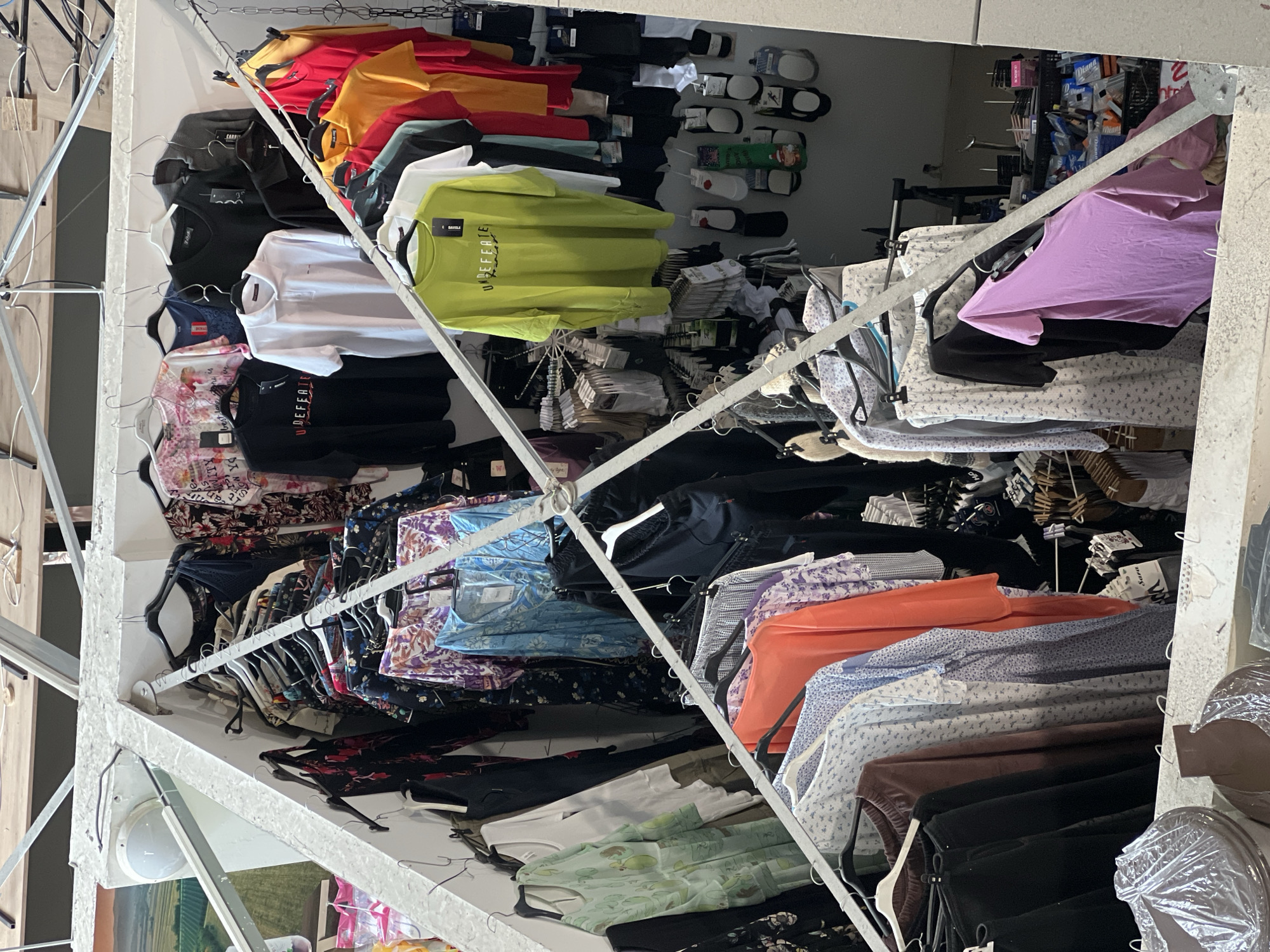 This indoor scene depicts a clothing store, possibly located in Palilula Urban Municipality, Serbia, filled with various garments hanging on racks. There are t-shirts in different colors (red, yellow, orange, green, white, black) at the upper left, and a mix of blouses, shirts, and other clothing items in various colors and patterns on racks below and to the right. Socks and other accessories are visible on shelves in the background. The racks are supported by a white, angular metal structure. The overall impression is of a well-stocked retail environment with a focus on apparel.