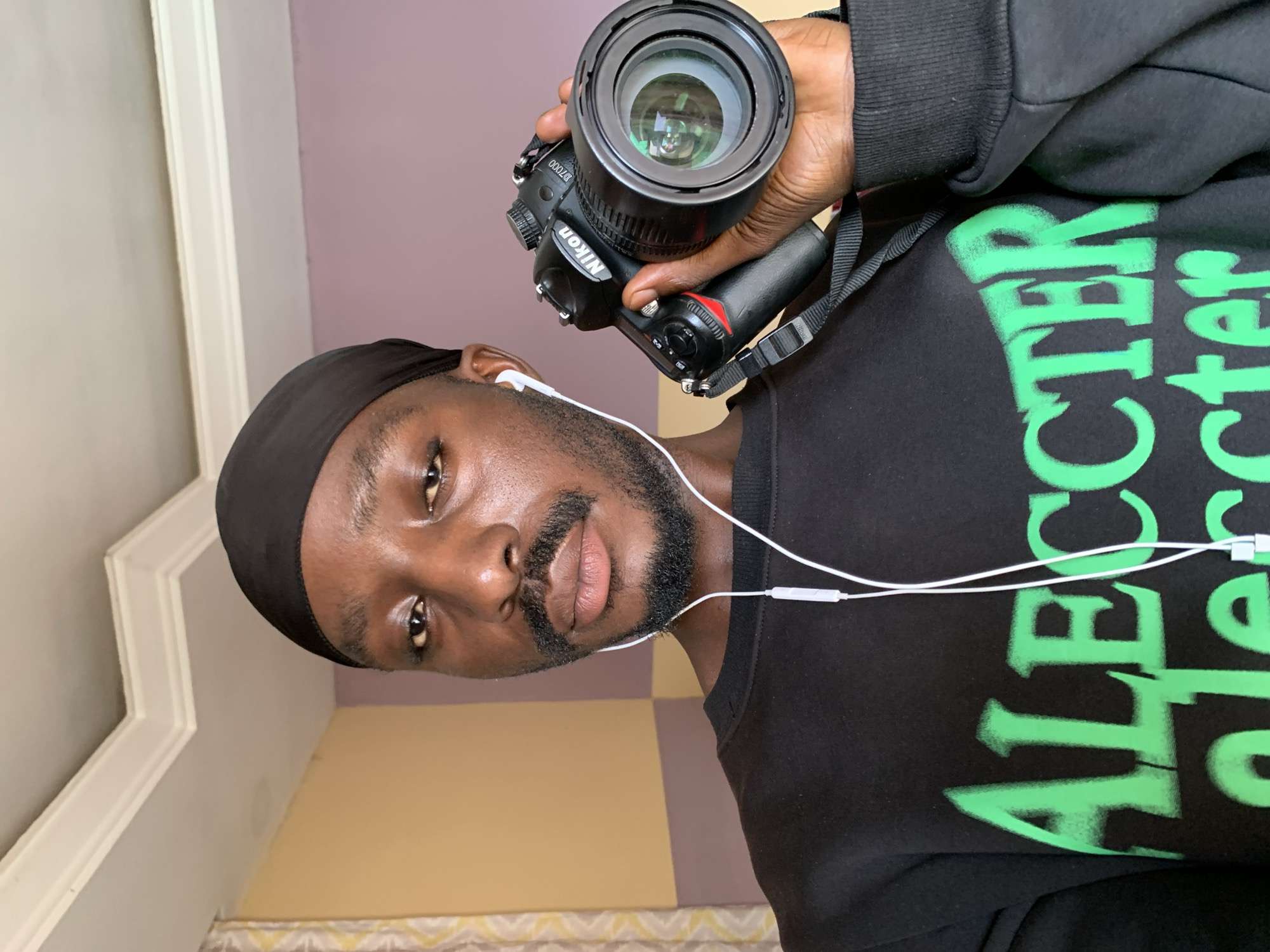 This is a selfie-style portrait of a Black man holding a Nikon D7000 camera. He is wearing a black headscarf, a black sweatshirt with green text that appears to read "ALECTER" and "STER," and white earbuds. The man has a beard and is looking towards the camera with a neutral expression.
The background suggests an indoor setting. To the left, there is a white architectural element, possibly a door frame or molding, with a textured ceiling above. To the right, the wall is painted in two distinct colors: a soft purple and a pale yellow, with a patterned curtain visible at the bottom. The lighting appears to be artificial, coming from above.
The man is holding the camera up with his right hand, and the camera strap is visible around his neck and resting on his shoulder. The lens of the camera is pointed towards the viewer. The overall composition suggests the man is in the process of taking a photograph of himself. The image appears to be taken during the daytime, but the indoor setting makes it difficult to determine the time of day or weather conditions. There are no specific details to pinpoint the exact location beyond the provided context of Jos, Nigeria.
