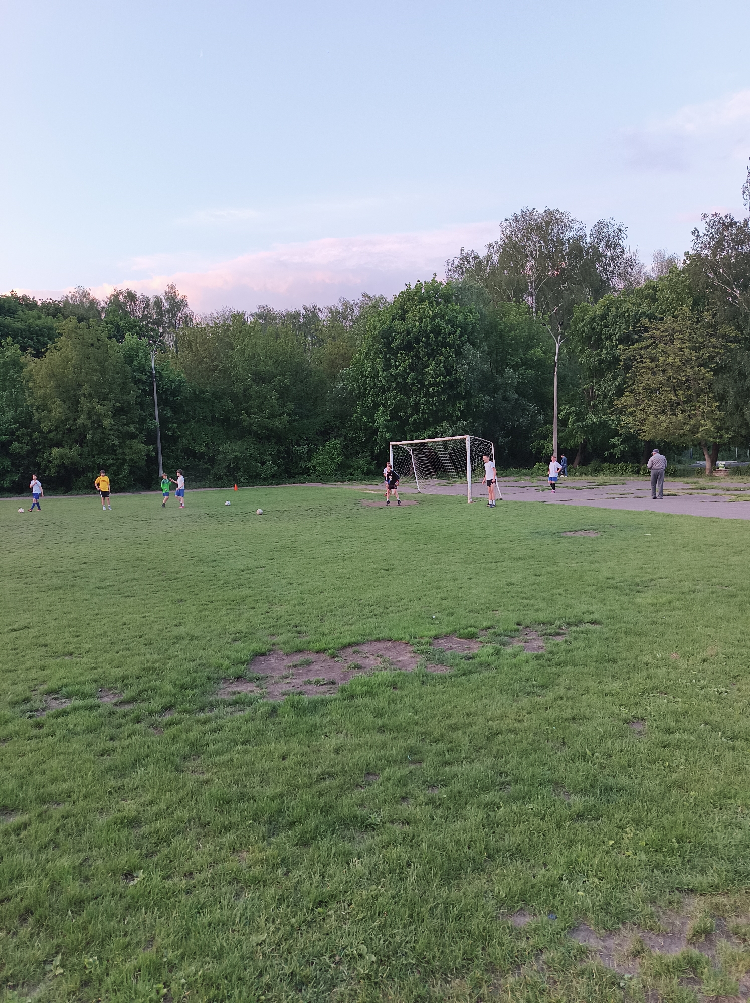 The image shows a group of people playing soccer on a grassy field, likely in Ternopil, Ukraine, based on prior data. There are several individuals dispersed across the field, some near a soccer goal. The terrain is primarily green grass, with some bare patches of dirt. In the background are trees and a light-colored sky with some clouds. One person is walking on a concrete path adjacent to the field. A soccer net is present in the center of the image.