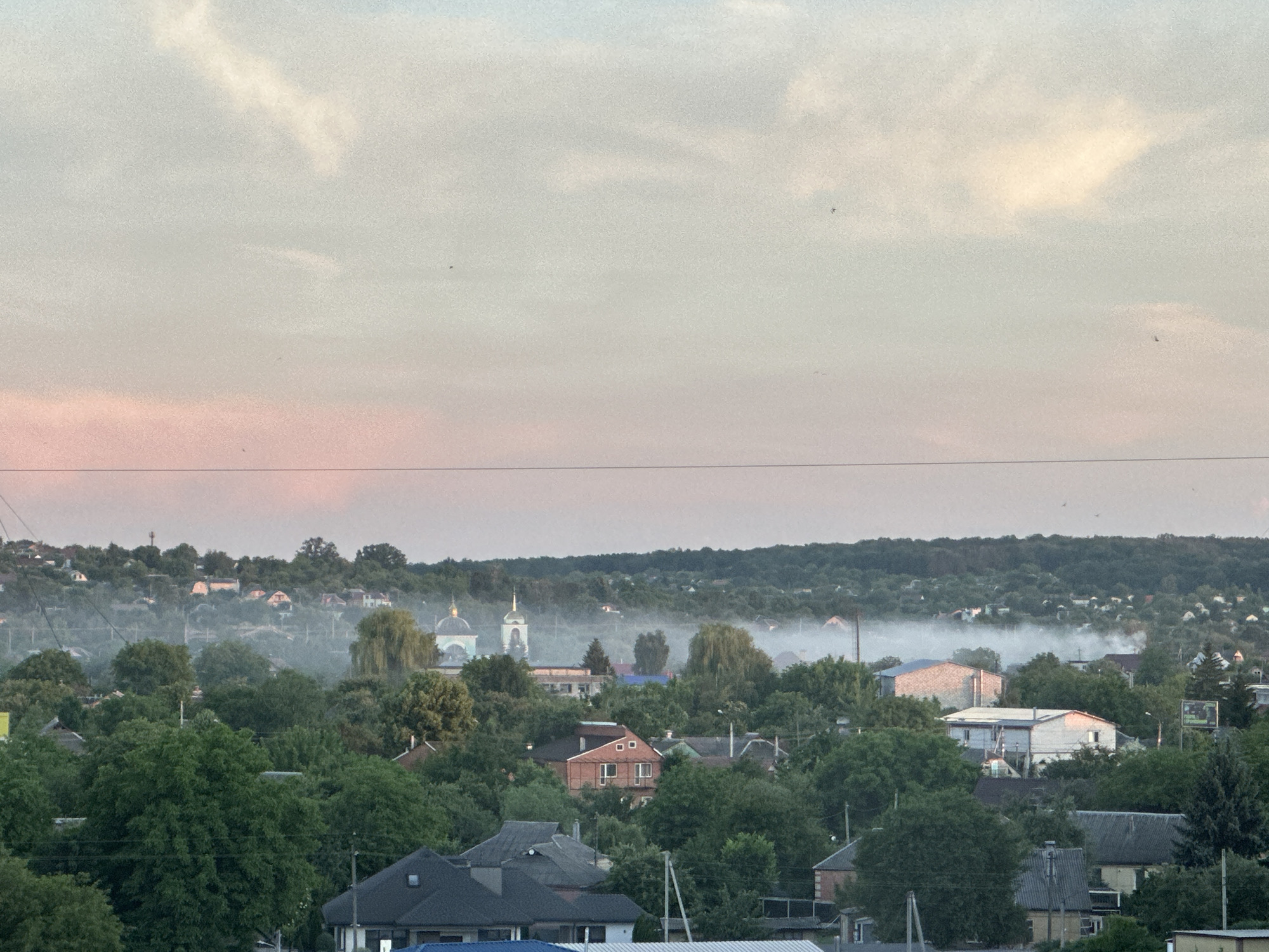 The image shows an aerial view of a semi-rural settlement at dawn. The sky is a gradient of pale blue to pink, with scattered clouds. In the foreground, several houses with dark roofs are visible, surrounded by lush green trees. A church with a blue dome and a tall steeple is located in the mid-ground, along with other buildings. A thin layer of mist or smoke hovers over the landscape, particularly in the area around the church and further into the distance. The distant hills are covered with dense forest. The scene conveys a sense of tranquility and the start of a new day in a quiet community.